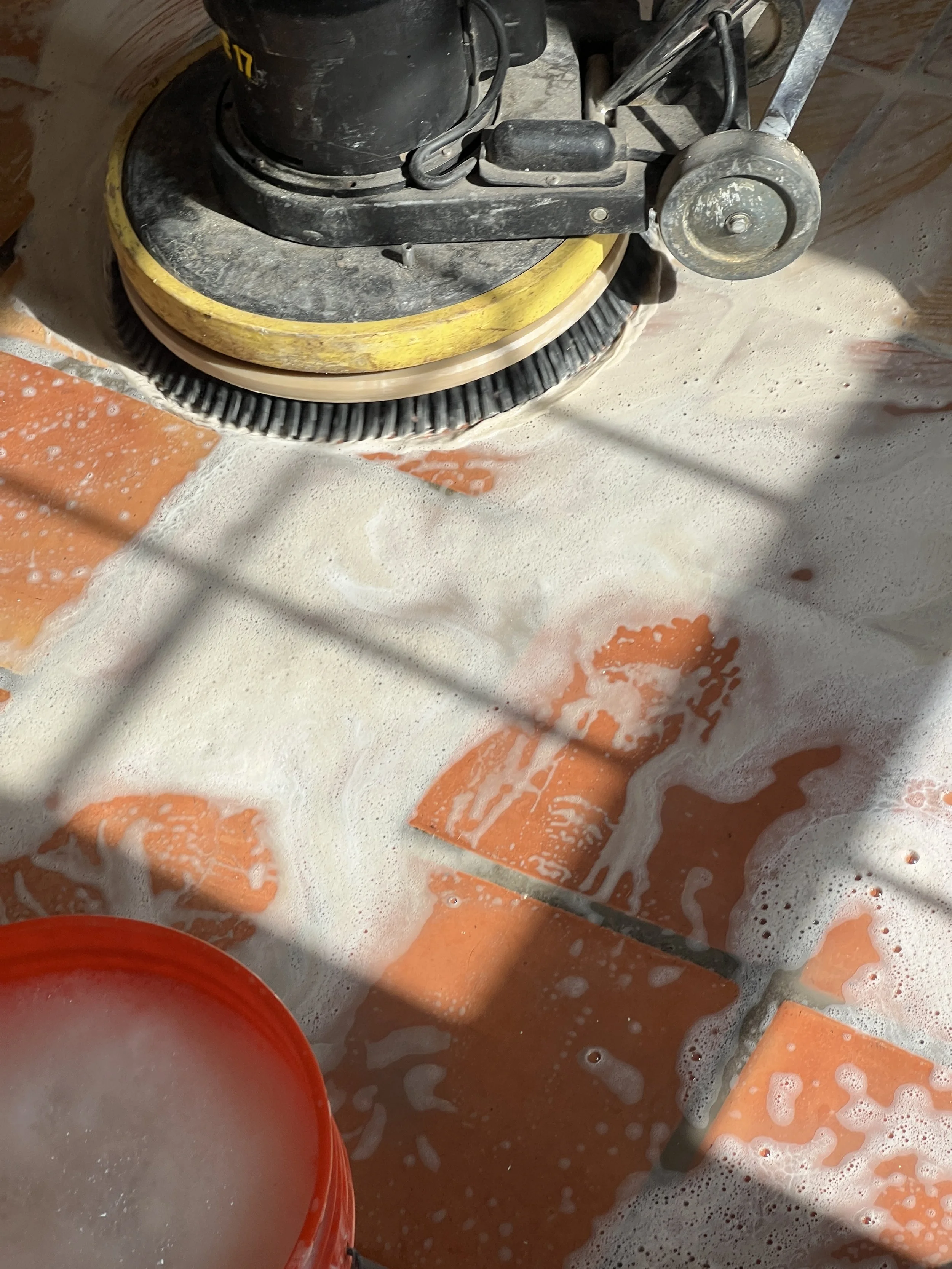 A floor with saltillo tiles and grout is being cleaned with a floor buffer that has a yellow and black base. Soap suds are visible on the tiles, and a red bucket is partially seen in the lower left corner.