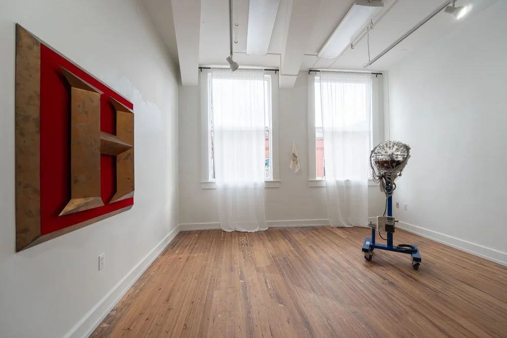 Empty room with hardwood floors, white walls, and two large windows with sheer white curtains. A red and wood wall decoration is on the left. A movable work lamp with a plastic cover is in the right corner.