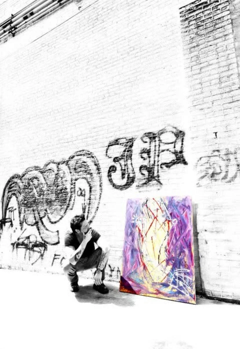 A person crouching in front of a colorful abstract painting leaning against a white brick wall with graffiti and graffiti text in a black-and-white photo.