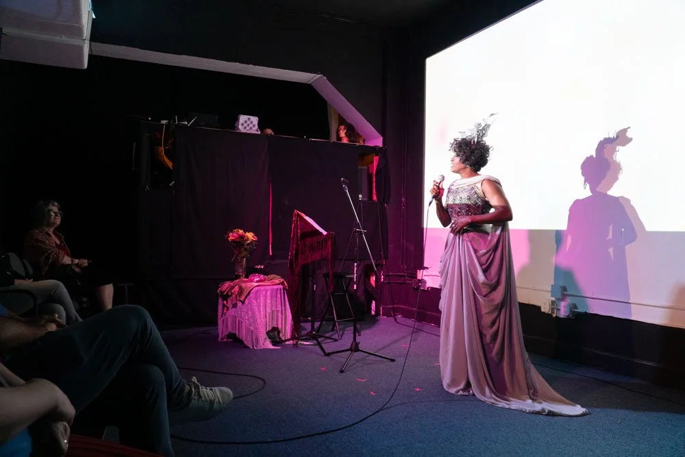 Performer in a purple gown singing into a microphone on stage while an audience watches.