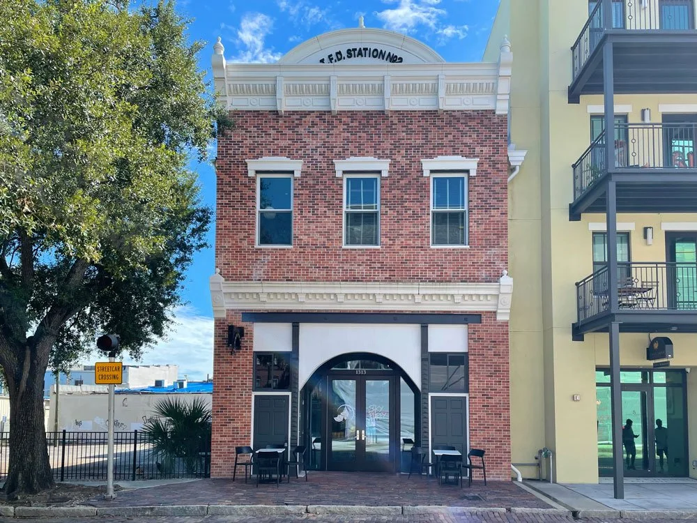A three-story building with a brick facade, white decorative trim, and a rounded arch entrance, situated between a large leafy tree on the left and a modern apartment building on the right, with a blue sky overhead.