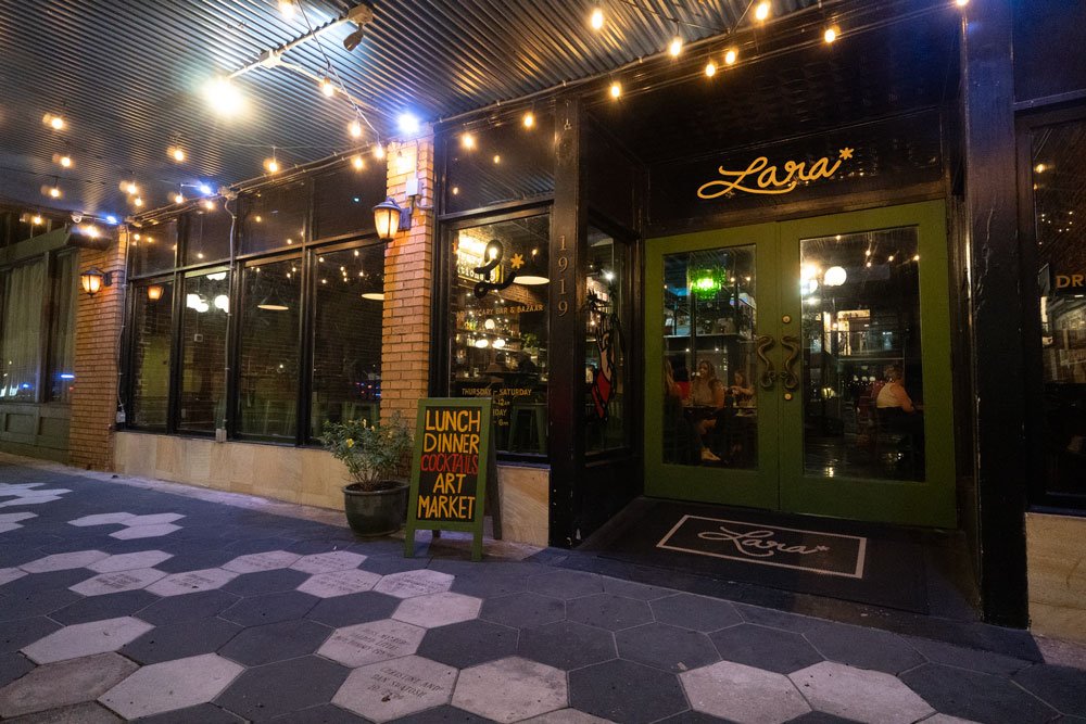 Night view of a restaurant named 'Lara' with green double doors, large window panels, and string lights above, located on a sidewalk with a chalkboard sign advertising lunch, dinner, cocktails, art, and market.