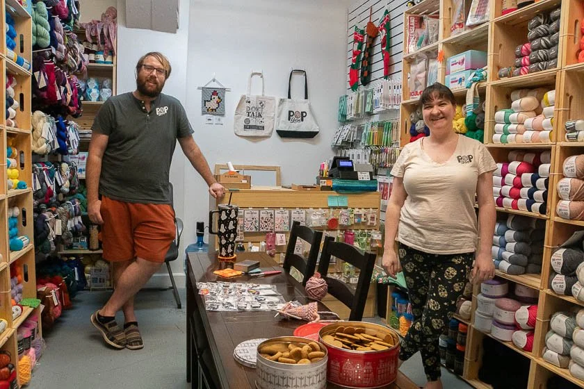 Two people standing inside a craft store surrounded by yarn and craft supplies, with a table displaying small items and cookies.