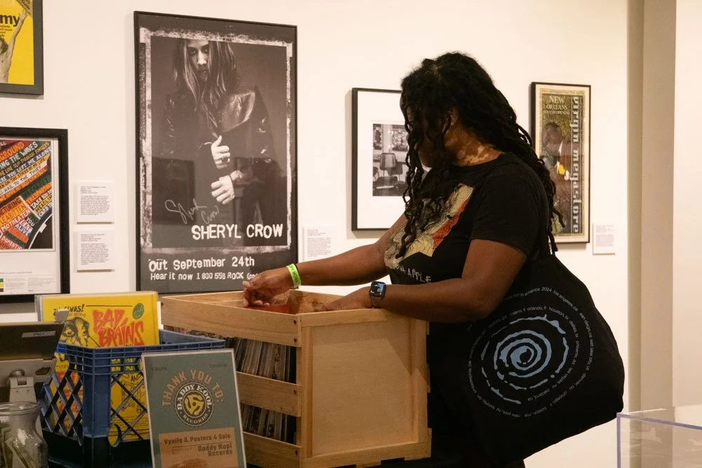A woman with long, curly hair and glasses looking into a wooden record crate in an art gallery.