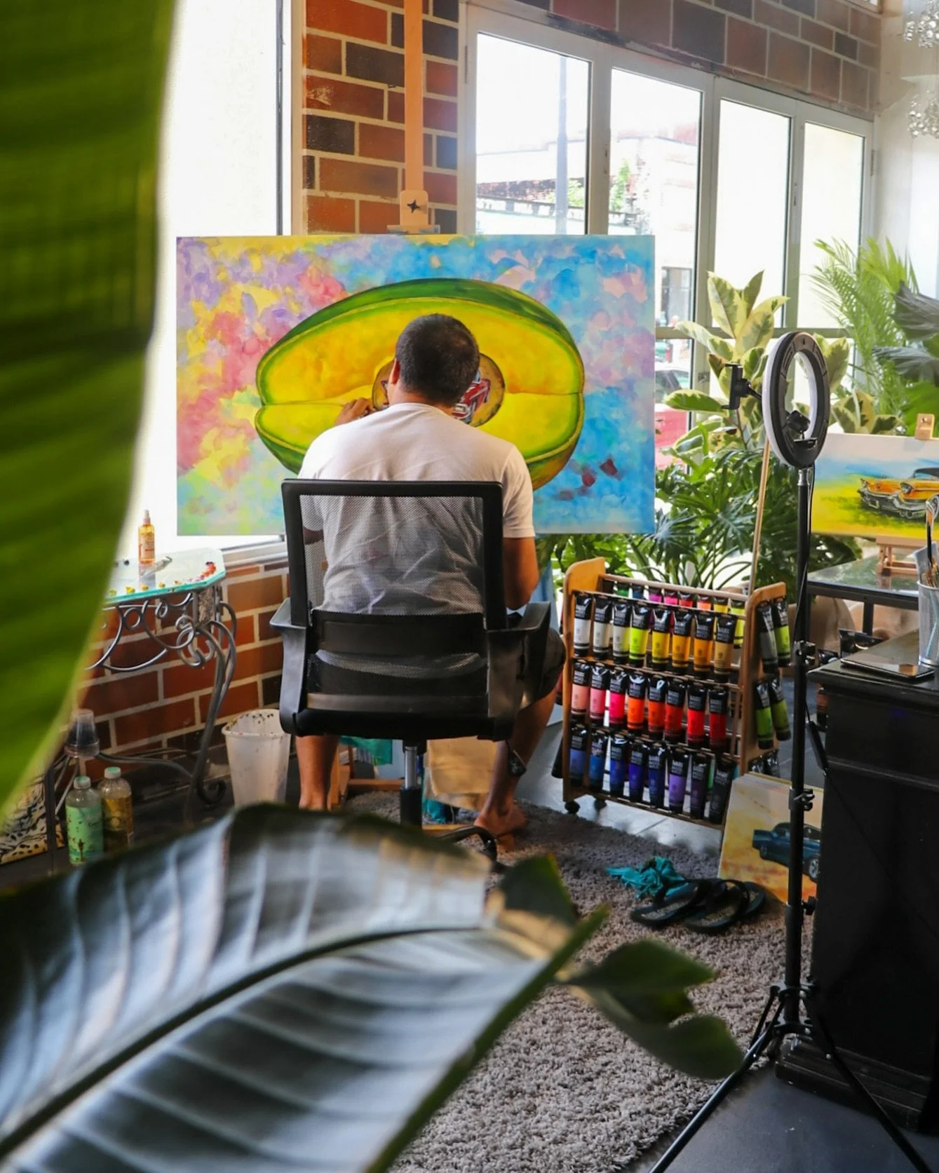 An artist painting a colorful canvas featuring a large avocado and a small red car, in a bright sunlit studio with plants and art supplies.