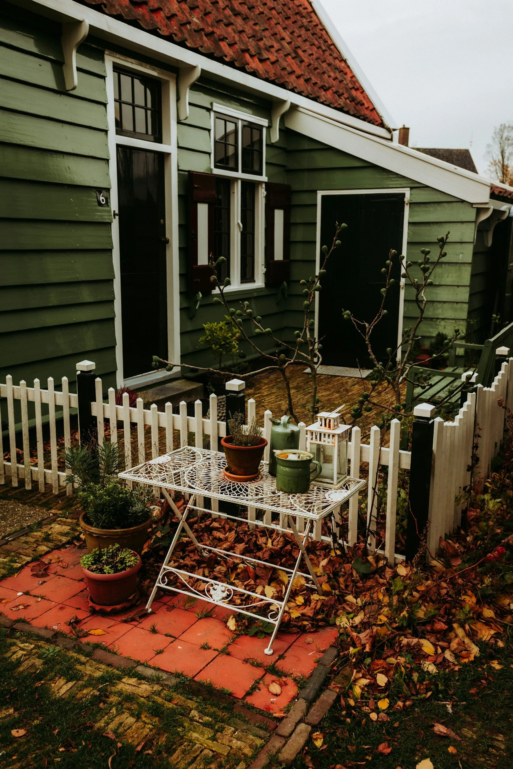 A small garden patio in front of a green house with white trim and dark brown shutters. The patio has a white metal table with potted plants, a lantern, and garden tools. Fallen leaves surround the area, and a white picket fence encloses the space.