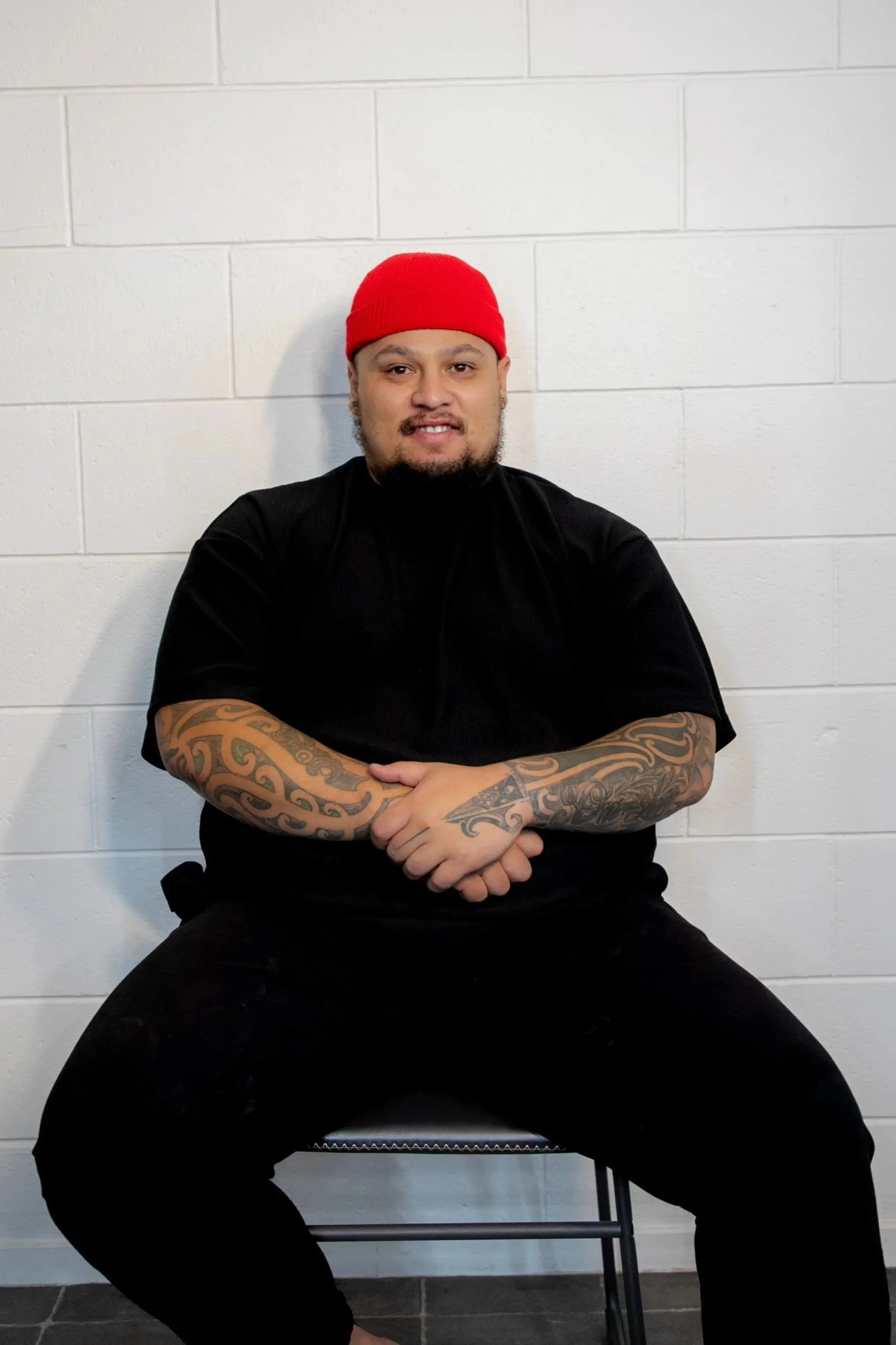 A Māori man with tattoos on his arms, wearing a red beanie, black shirt, and black pants, sitting cross-legged against a white brick wall. Māori Graphic Designer
