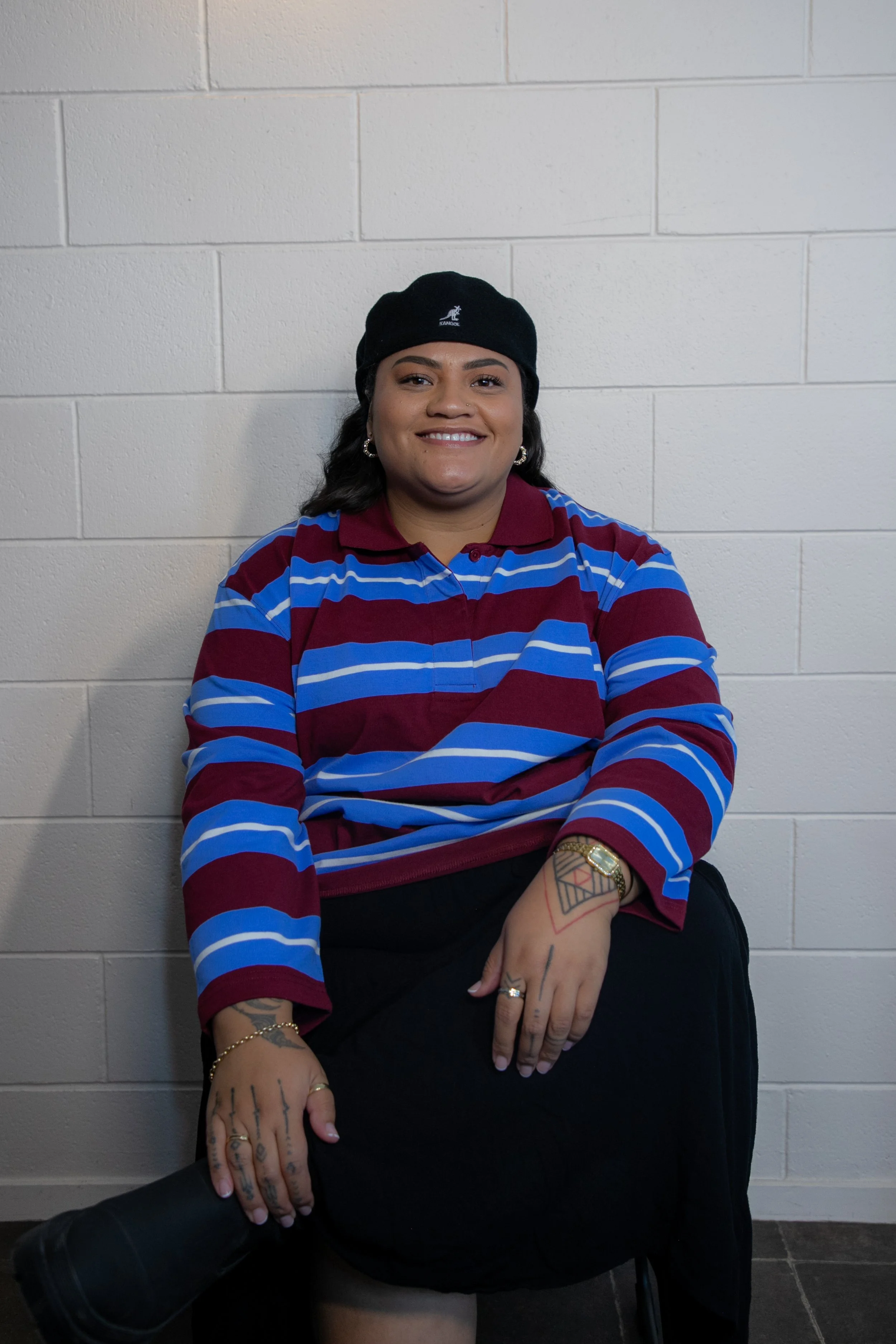 A Māori woman smiling, sitting against a white brick wall, wearing a black beanie, a striped maroon and blue shirt, with tattoos on her hands, and jewelry including a watch, ring, and bracelets, Māori Graphic Designer