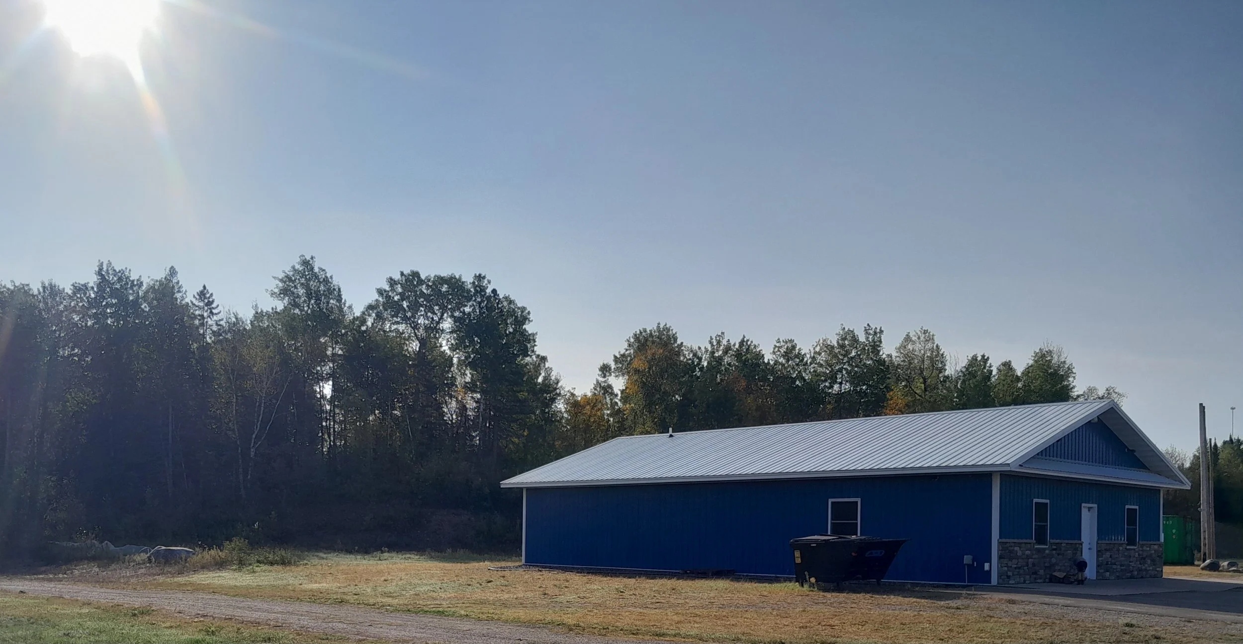 A blue building with a silver metal roof in front of a wooded area and a clear blue sky with the sun shining brightly.