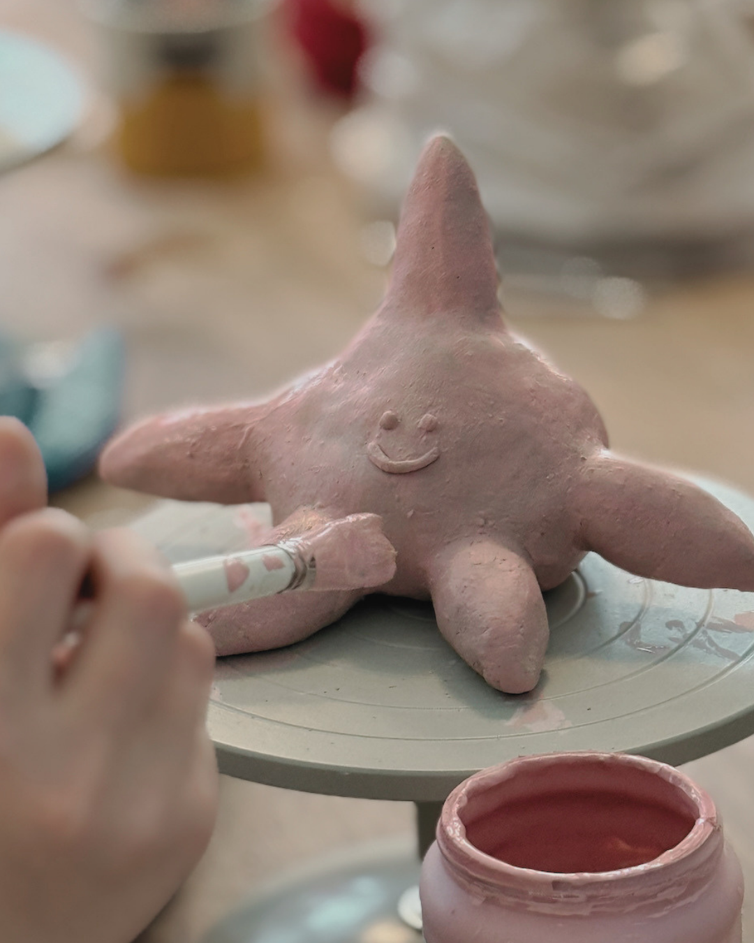 Eine rosa Tonfigur eines Glückskopfes mit einem lächelnden Gesicht und fünf Armen, die auf einem Töpferscheibe steht, während jemand mit einem Pinsel die Figur bemalt.