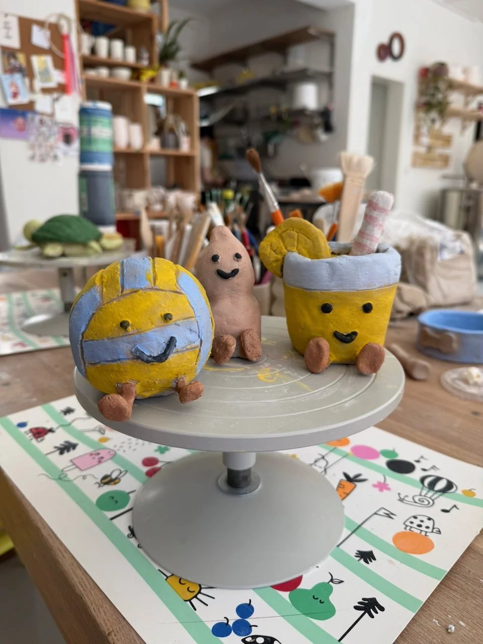 Bunte, lächelnde Keramikfiguren eines Wackelpuddings mit Stäbchen, eines Quallenschirms und eines Augapfels auf einem drehbaren Ständer in einer Kunstatelier-Umgebung.