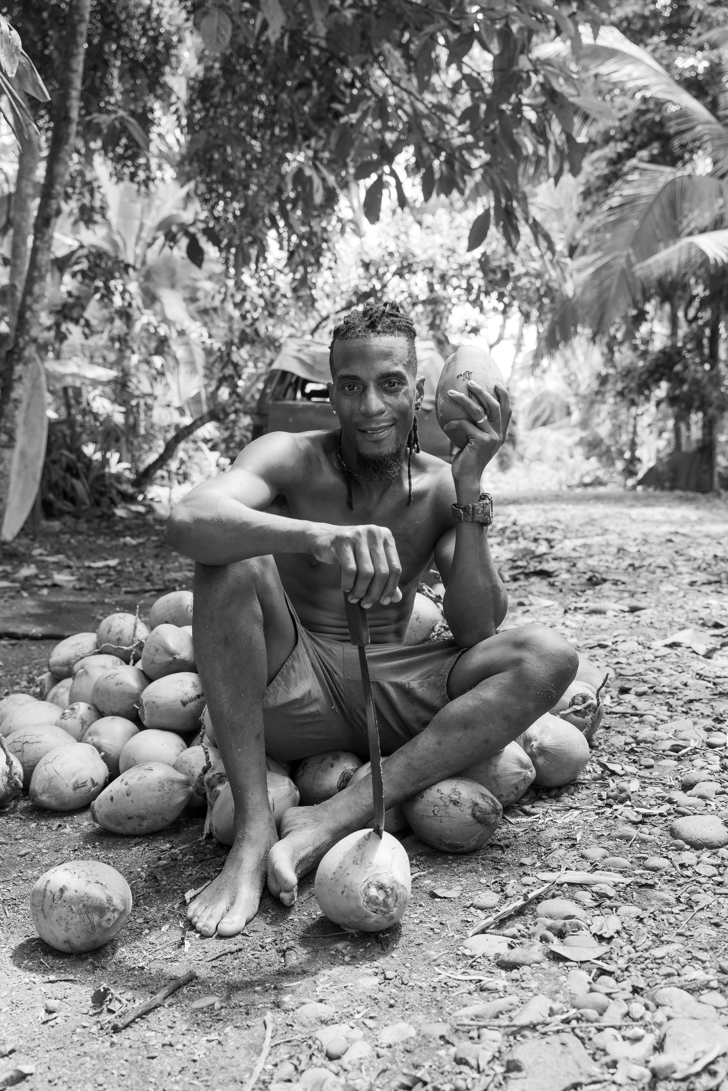 A man sitting barefoot on the ground surrounded by coconuts, holding a coconut in one hand and a knife in the other, with a jungle background.
