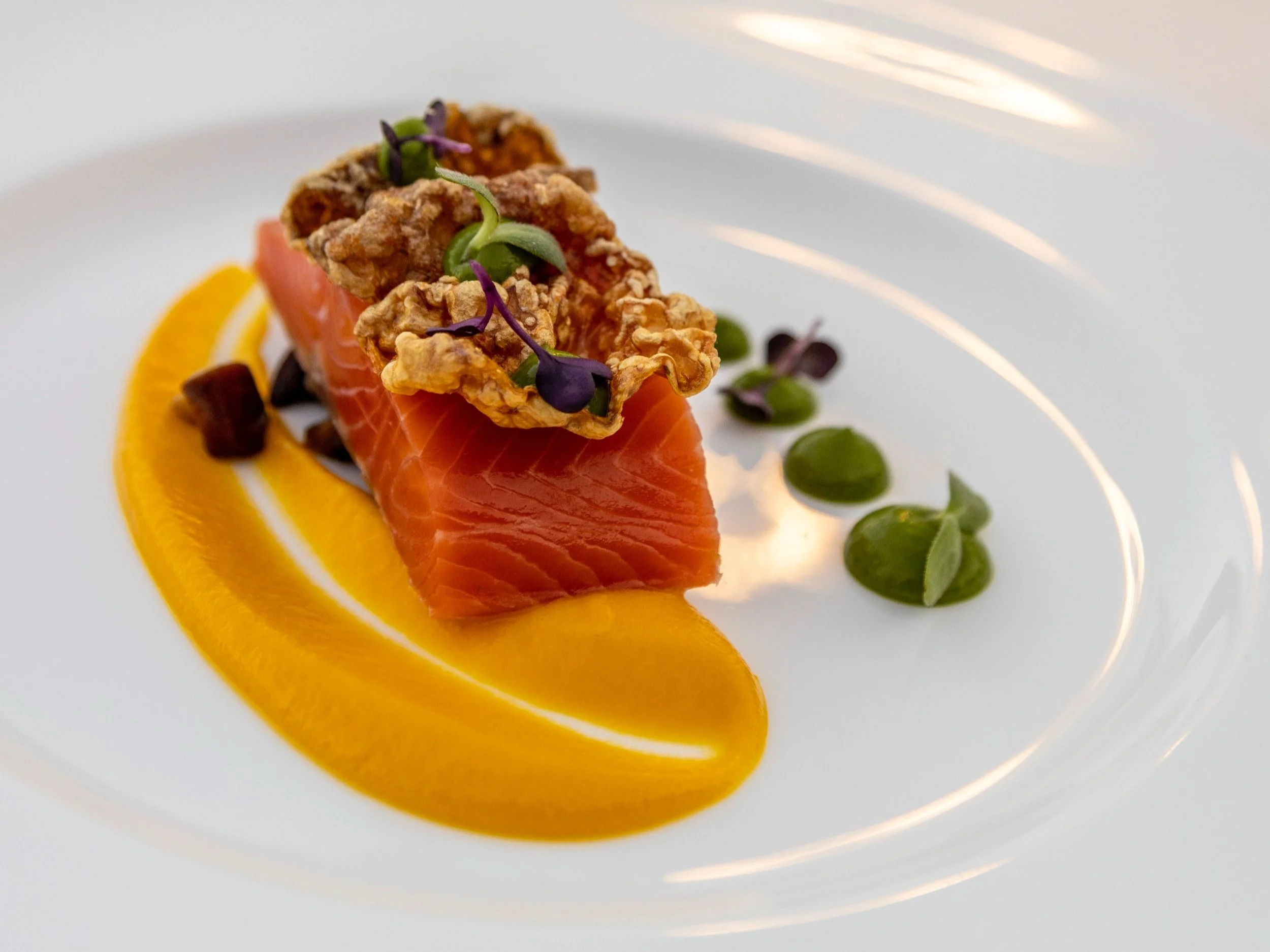 A dish of fresh salmon sashimi topped with crispy garnish and microgreens, served with slices of orange and green beans on a white plate.