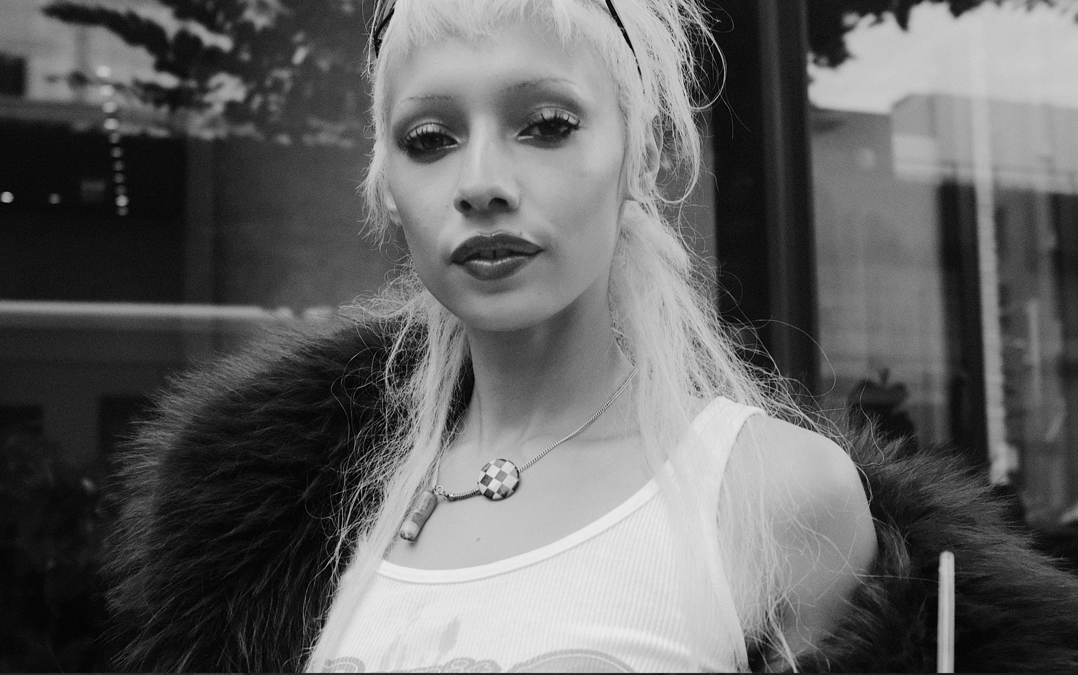 A split-screen black and white photo, with a woman with long, platinum blonde hair in the top half and a man in a suit and mask standing next to a black luxury car in the bottom half.