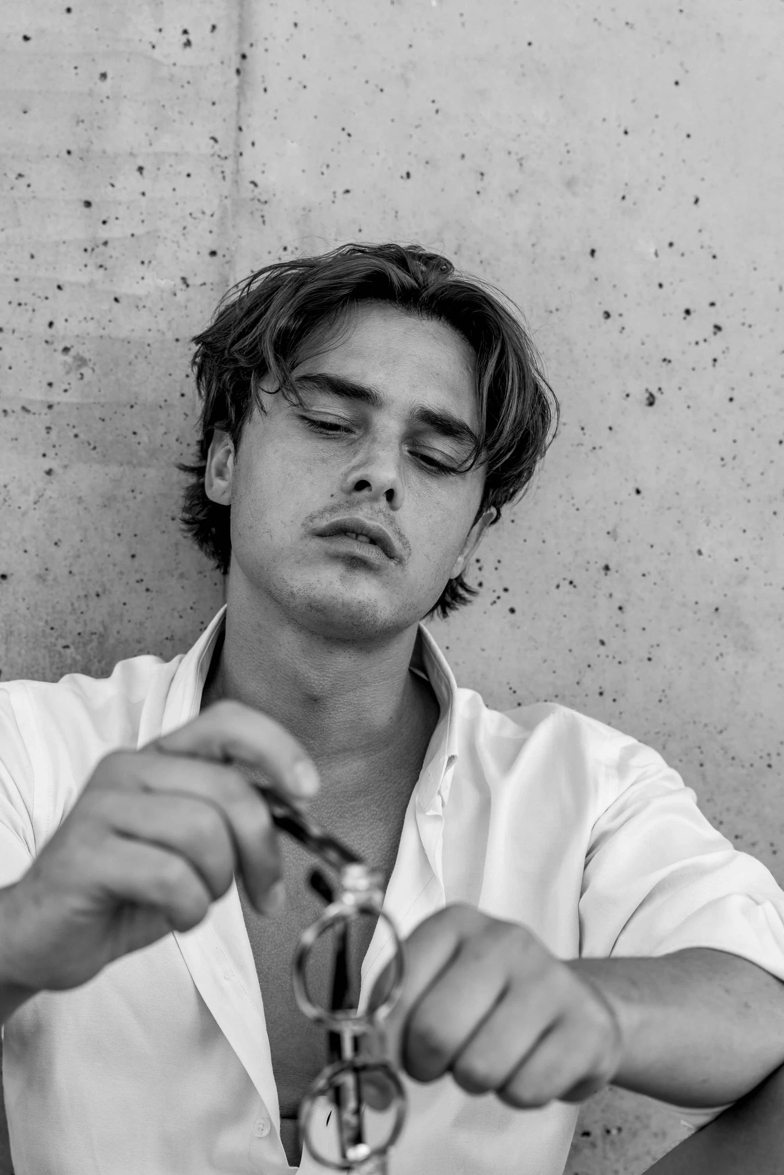 Black and white photo of a young man with messy hair, wearing an open collared shirt, looking down with a serious expression, holding a keychain or object in his hands, against a concrete wall background.