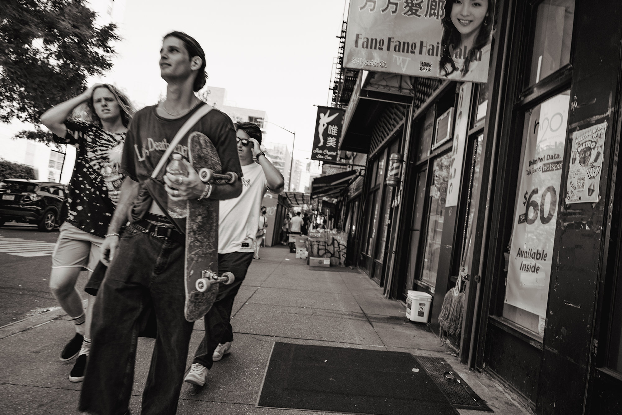 Street scene in an urban area with several pedestrians, including a young man holding a skateboard and water bottle, two women walking nearby, storefronts with signs, and parked cars in the background.