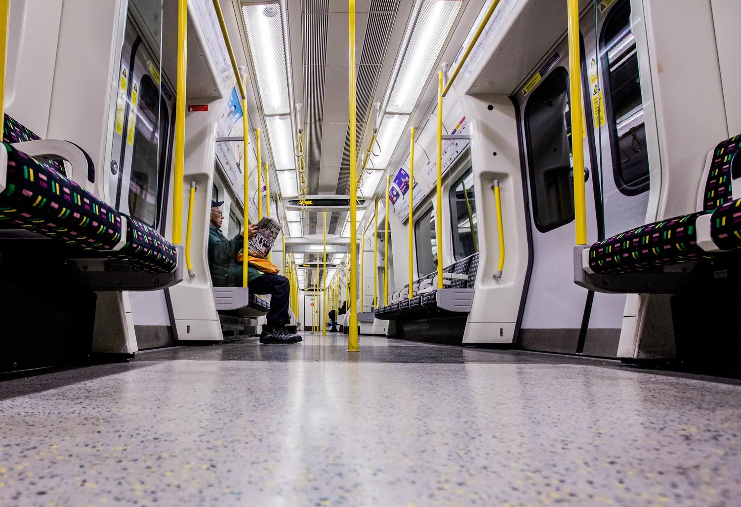 Inside a mostly empty subway train car with a low-angle view showing rows of black seats with colorful patterns, yellow poles, and one passenger reading a newspaper