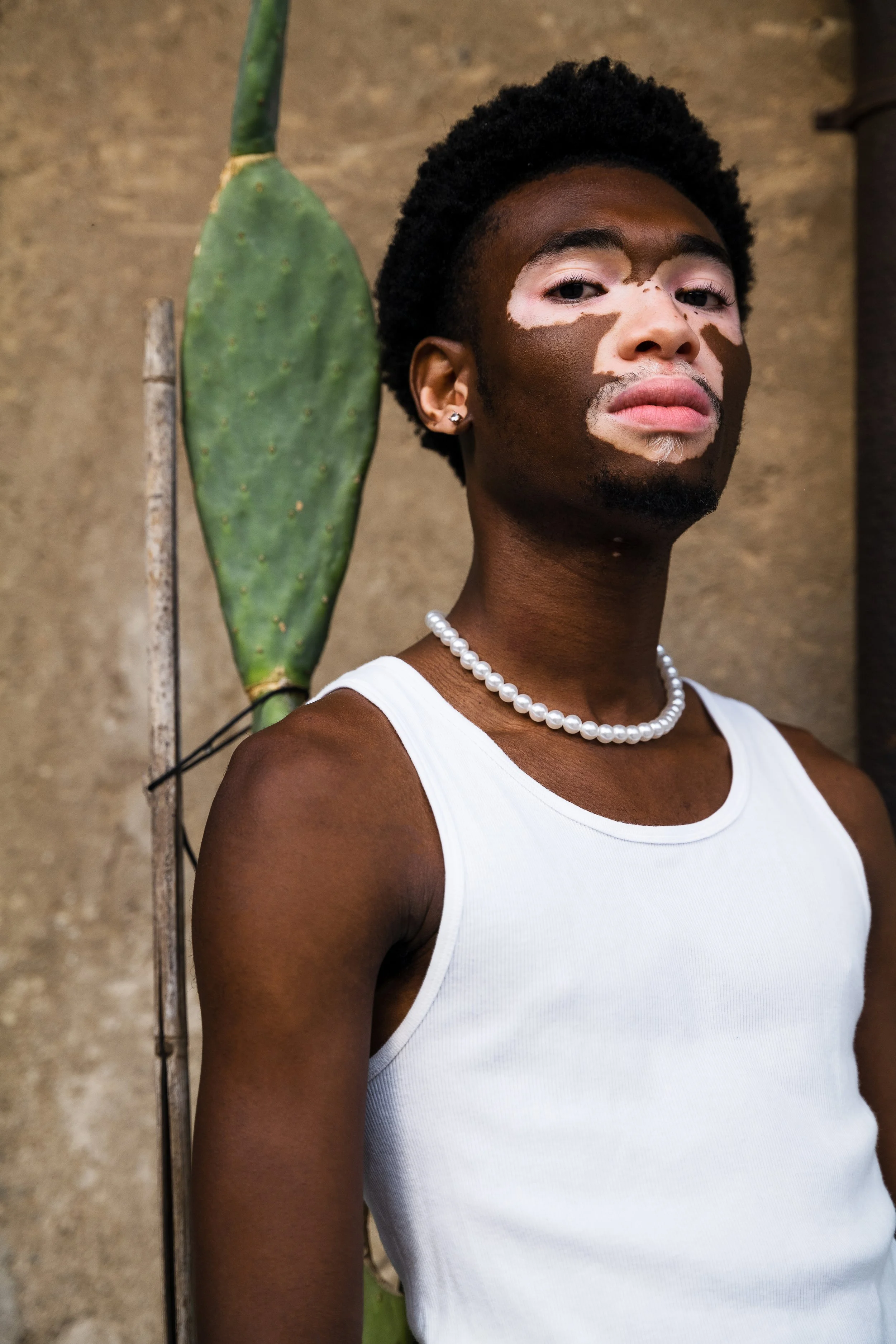 A person with vitiligo, wearing a white tank top and a pearl necklace, stands next to a cactus with a textured wall in the background.