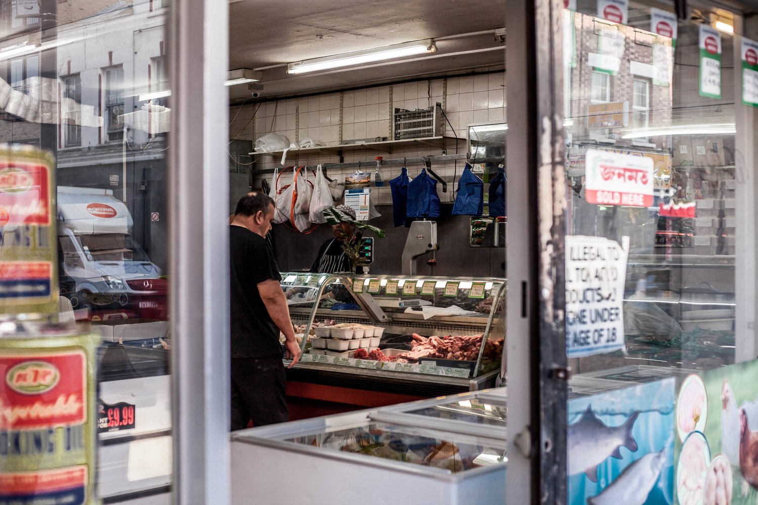 View through shop window showing a man inside a butcher shop with various meats on display, a butcher behind the counter, and blue aprons hanging on the wall.