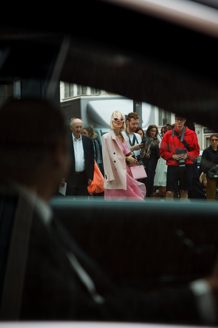 People waiting on a city sidewalk, viewed through the window of a parked car, including a woman in pink with sunglasses, a man using his phone, and a man in a red hoodie.