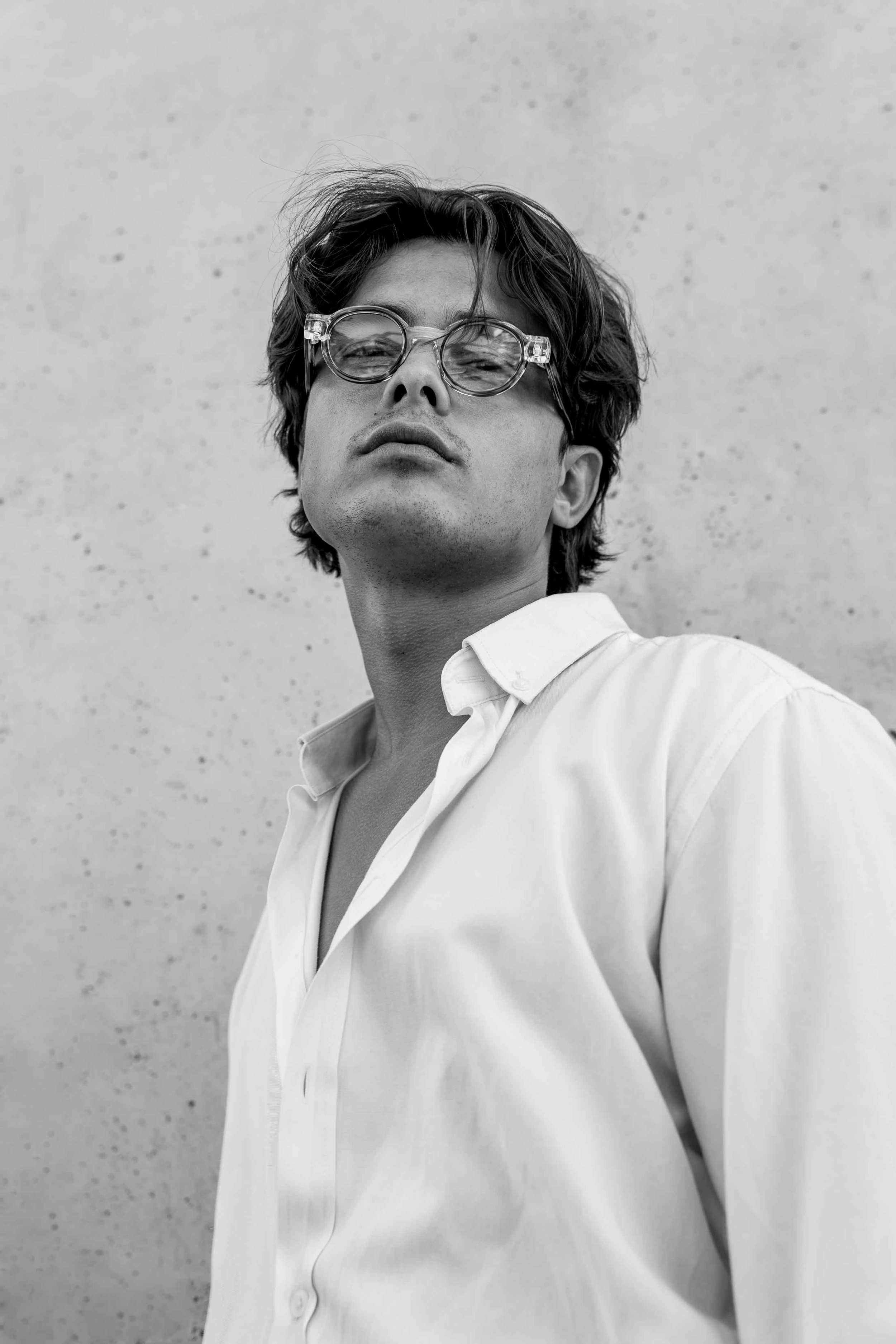 Black and white photo of a young man wearing glasses and a white shirt, looking confidently at the camera against a plain background.
