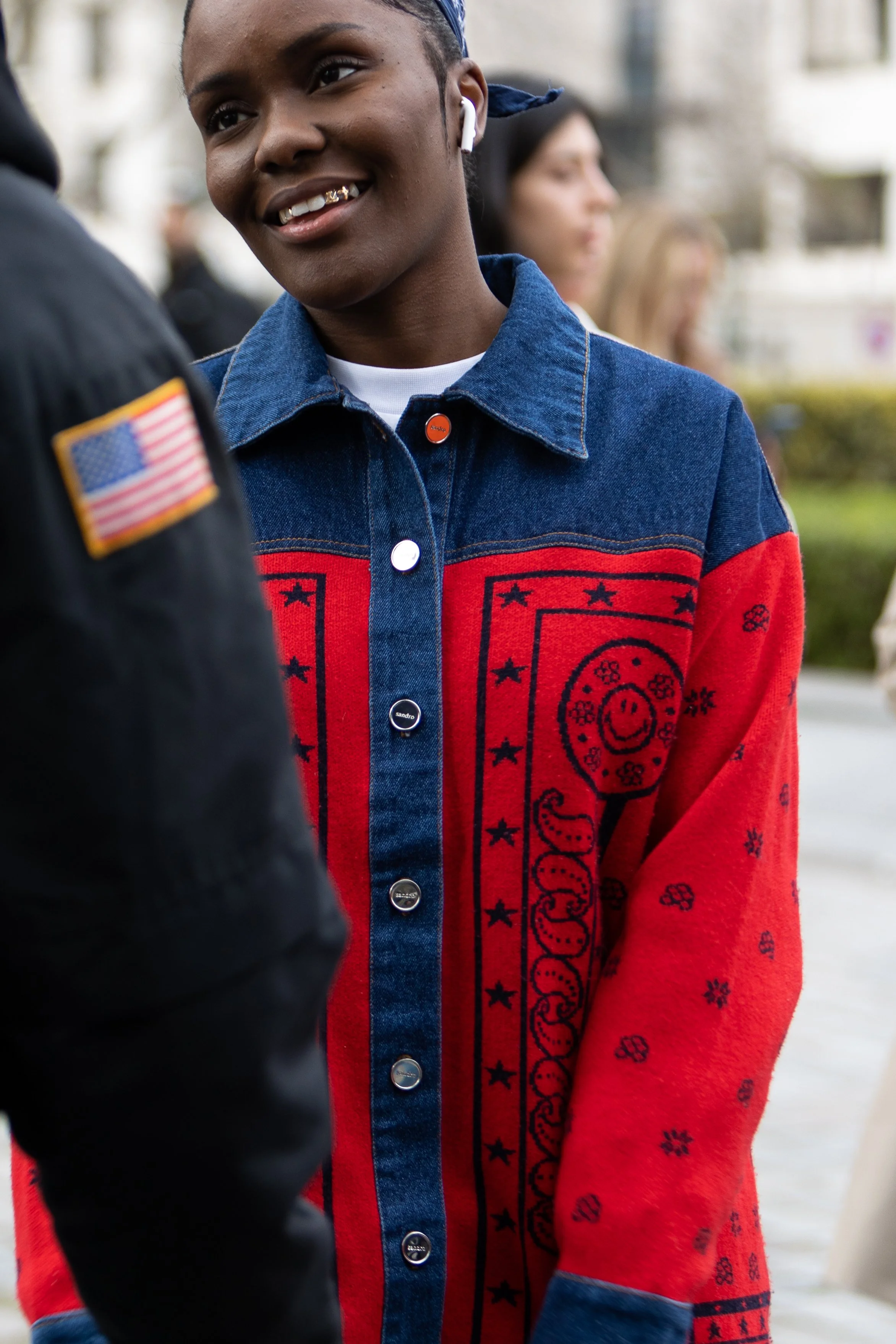 A person wearing a colorful patterned denim jacket talking to another person during an outdoor event, with some women blurred in the background.