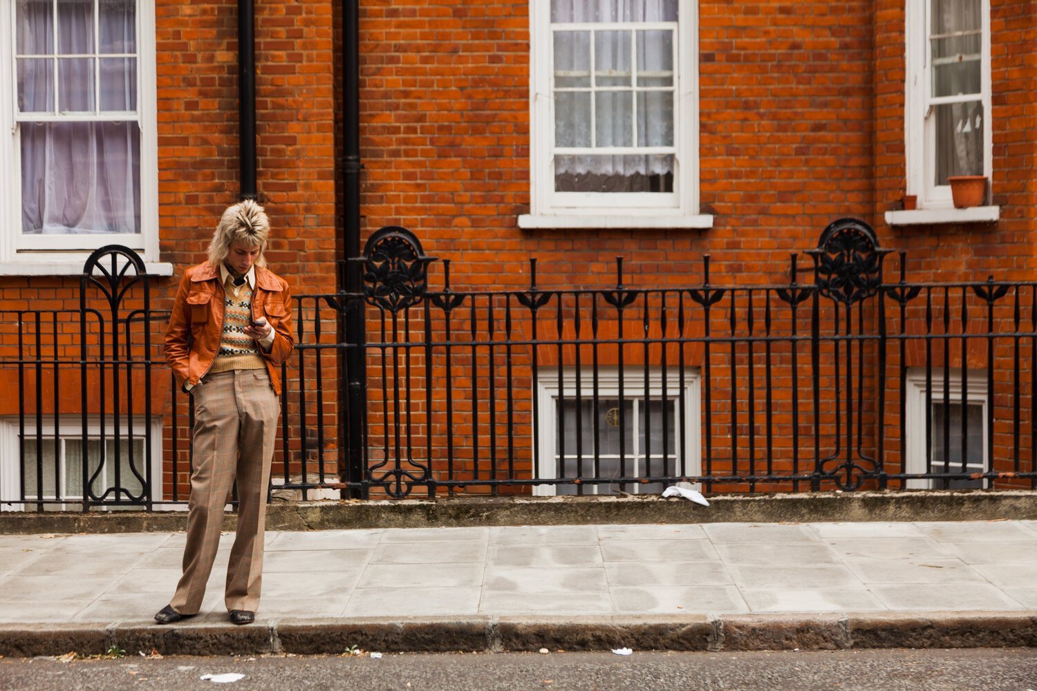 A woman with blonde hair wearing a brown jacket looking at her phone while standing on a sidewalk in front of a brick building with white window frames and a black metal fence.