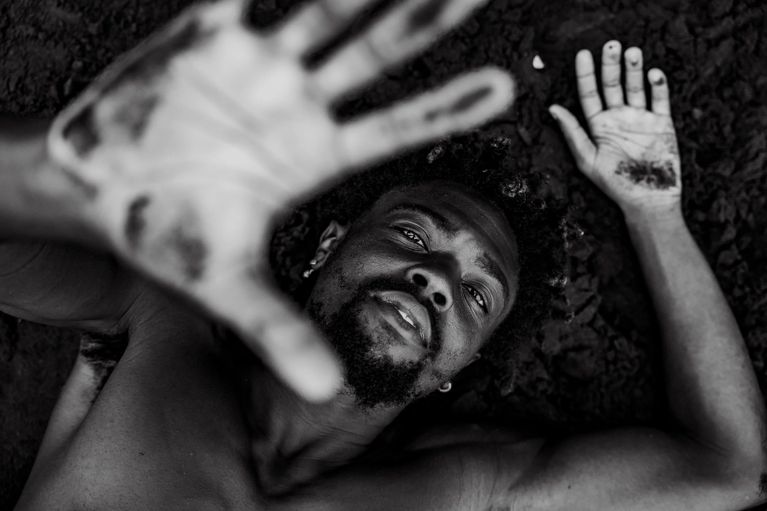 A shirtless man with a beard and curly hair is lying on dark soil, reaching towards the camera with his hand, while looking up. The image is in black and white.