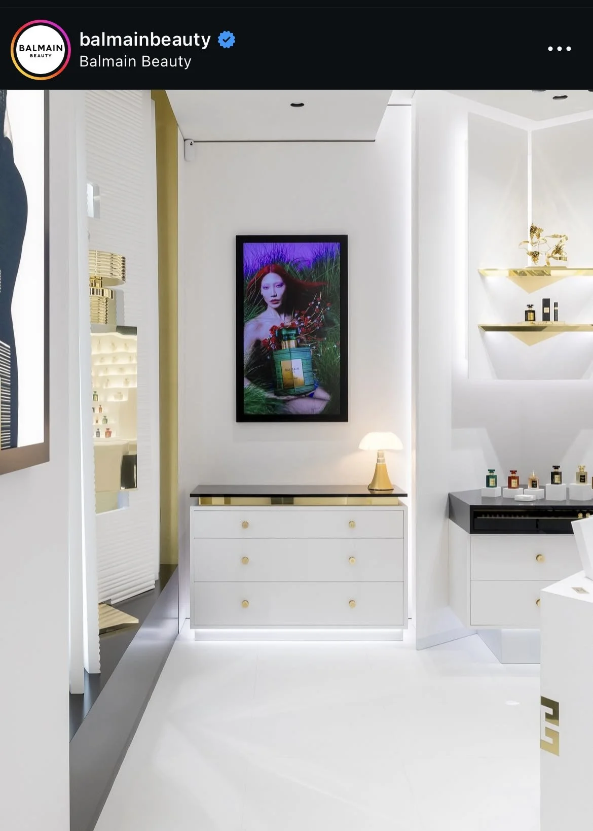 Interior of a modern beauty boutique with white walls, a digital screen featuring a woman, a white dresser with a lamp, gold shelving with beauty products, and sleek design elements.