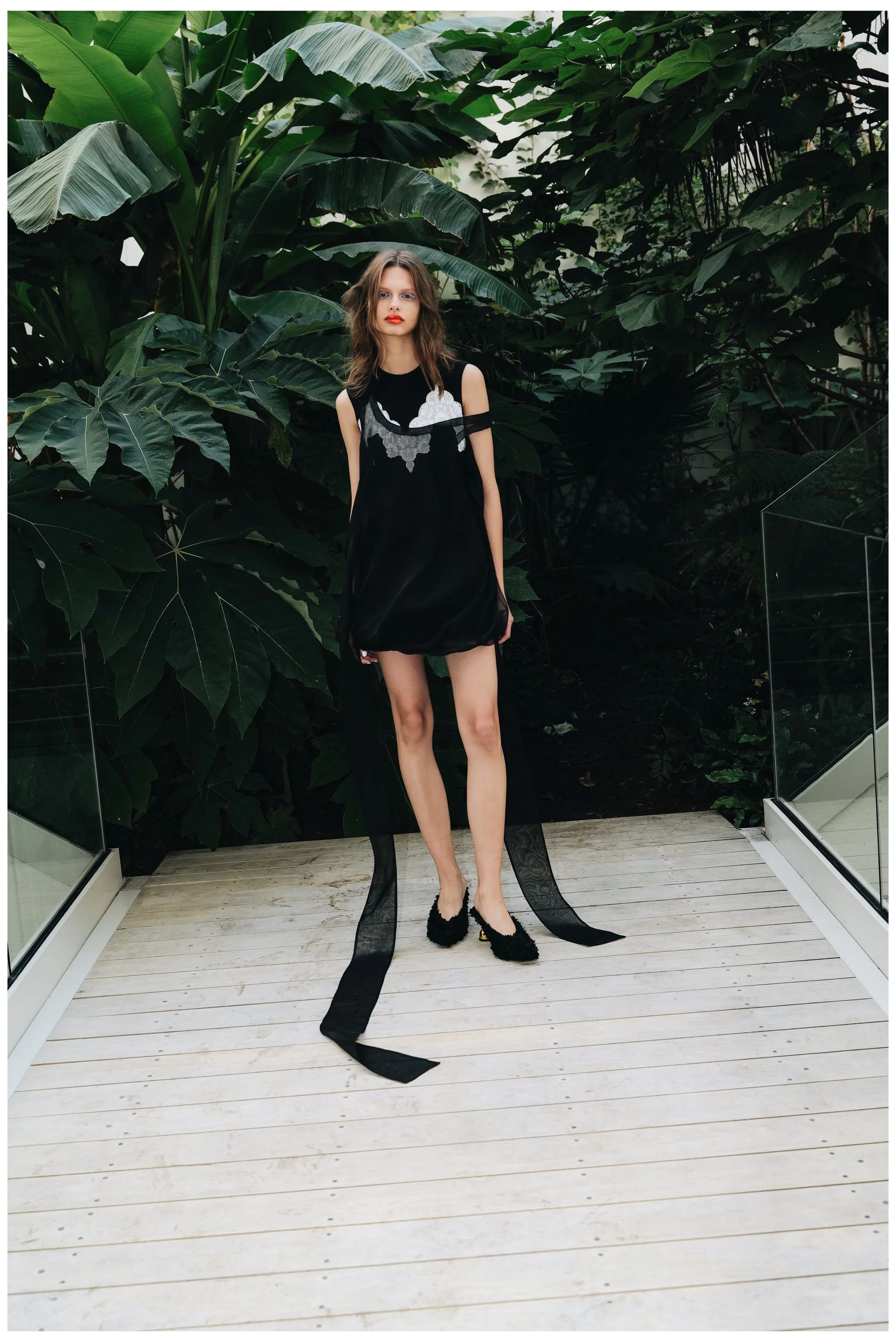 A young woman with shoulder-length brown hair wearing a black dress with cut-out shoulders and white embroidered details, standing on a light wooden deck with lush green tropical plants in the background.