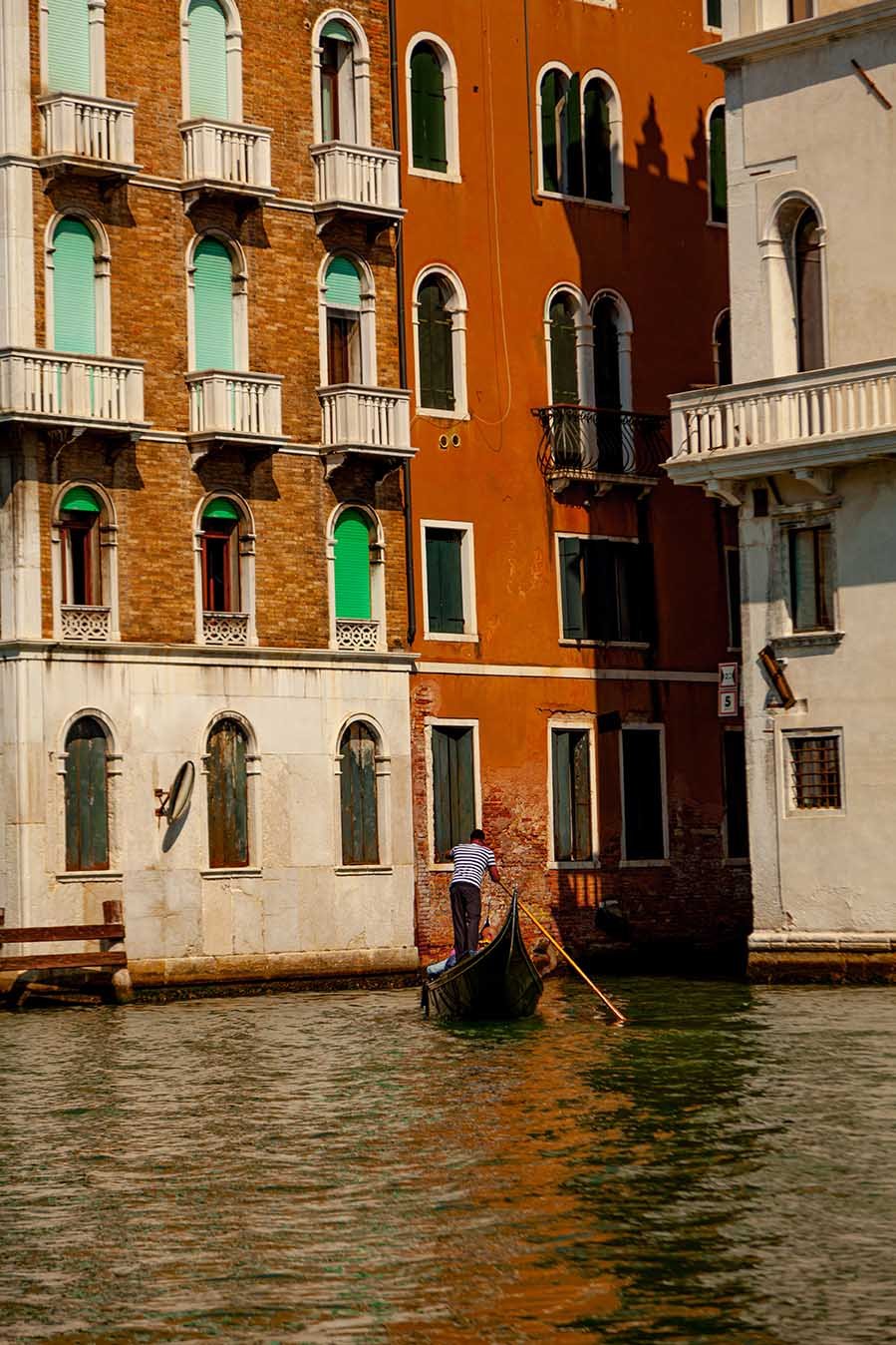 A gondolier in a striped shirt paddling a gondola in a canal alongside colorful old buildings with arched windows and small balconies in Venice, Italy.