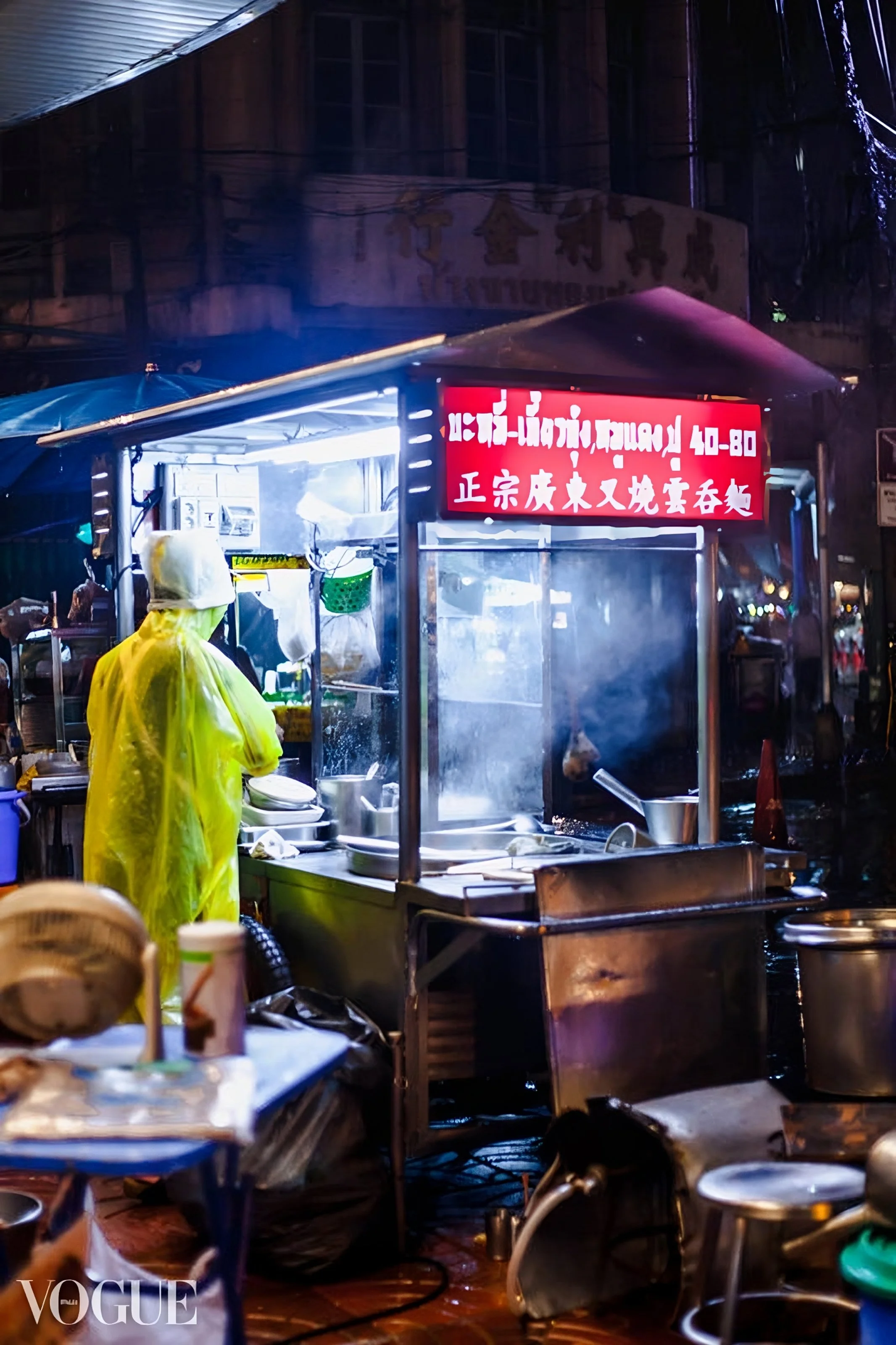 Night scene of a street food stall with a vendor wearing a yellow raincoat and hood, cooking in a steam-filled environment. The stall has a red illuminated sign with Chinese characters, and various cooking utensils and containers are visible, suggest