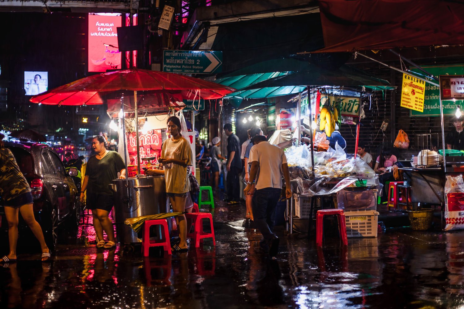 Night scene of a busy street market with several street vendors under umbrellas, illuminated by streetlights and neon signs, with people walking and shopping in the rain on a wet street.