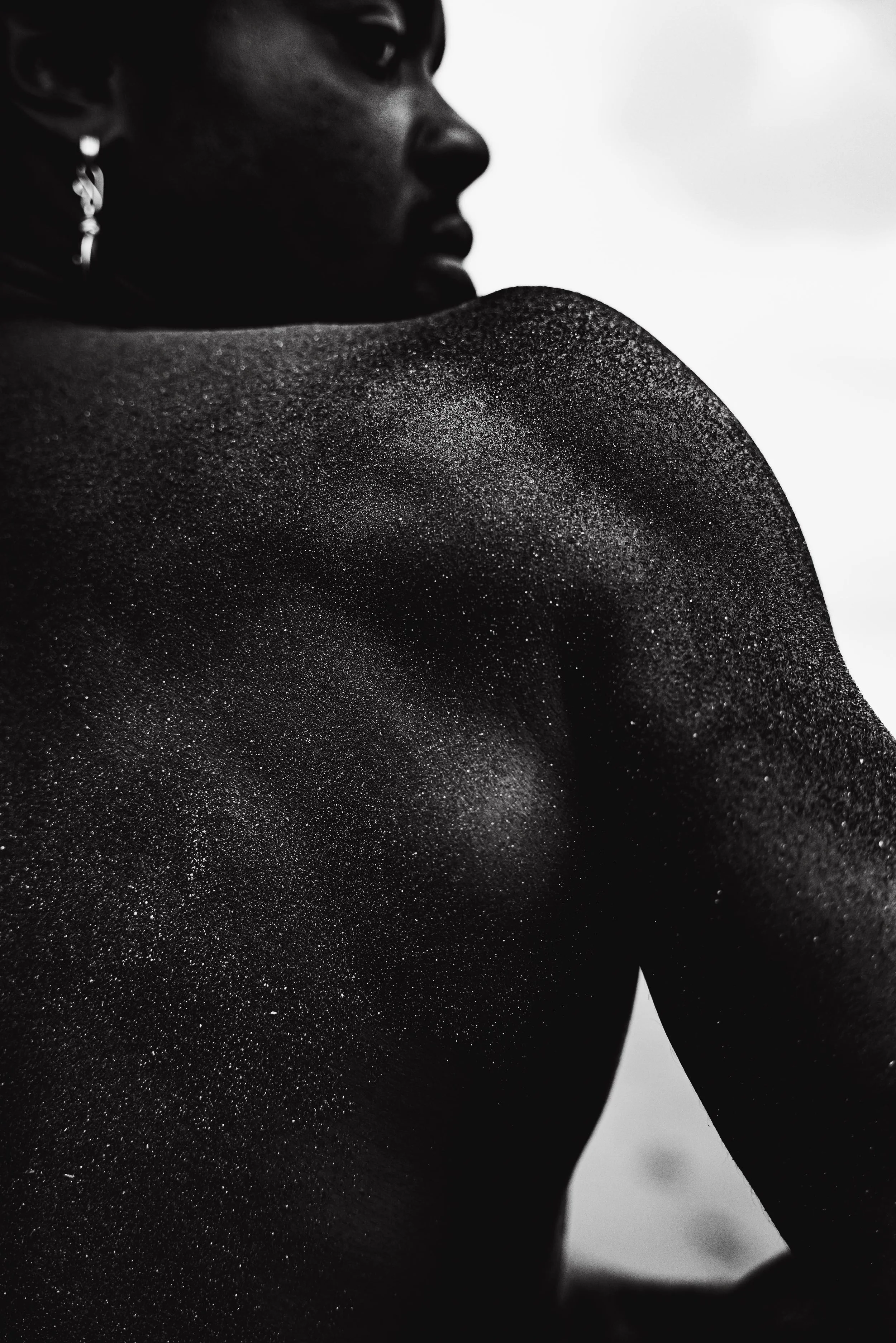 Close-up black and white photograph of a woman's shoulder and face, with water droplets on her skin, looking sideways.