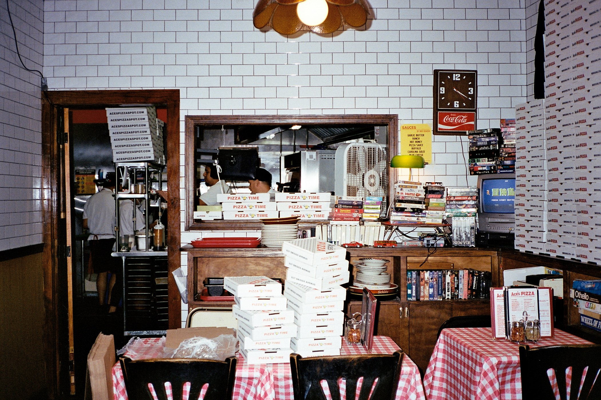 Interior of a pizzeria with shelves of pizza boxes, books, and a TV, alongside tables with red and white checkered tablecloths.