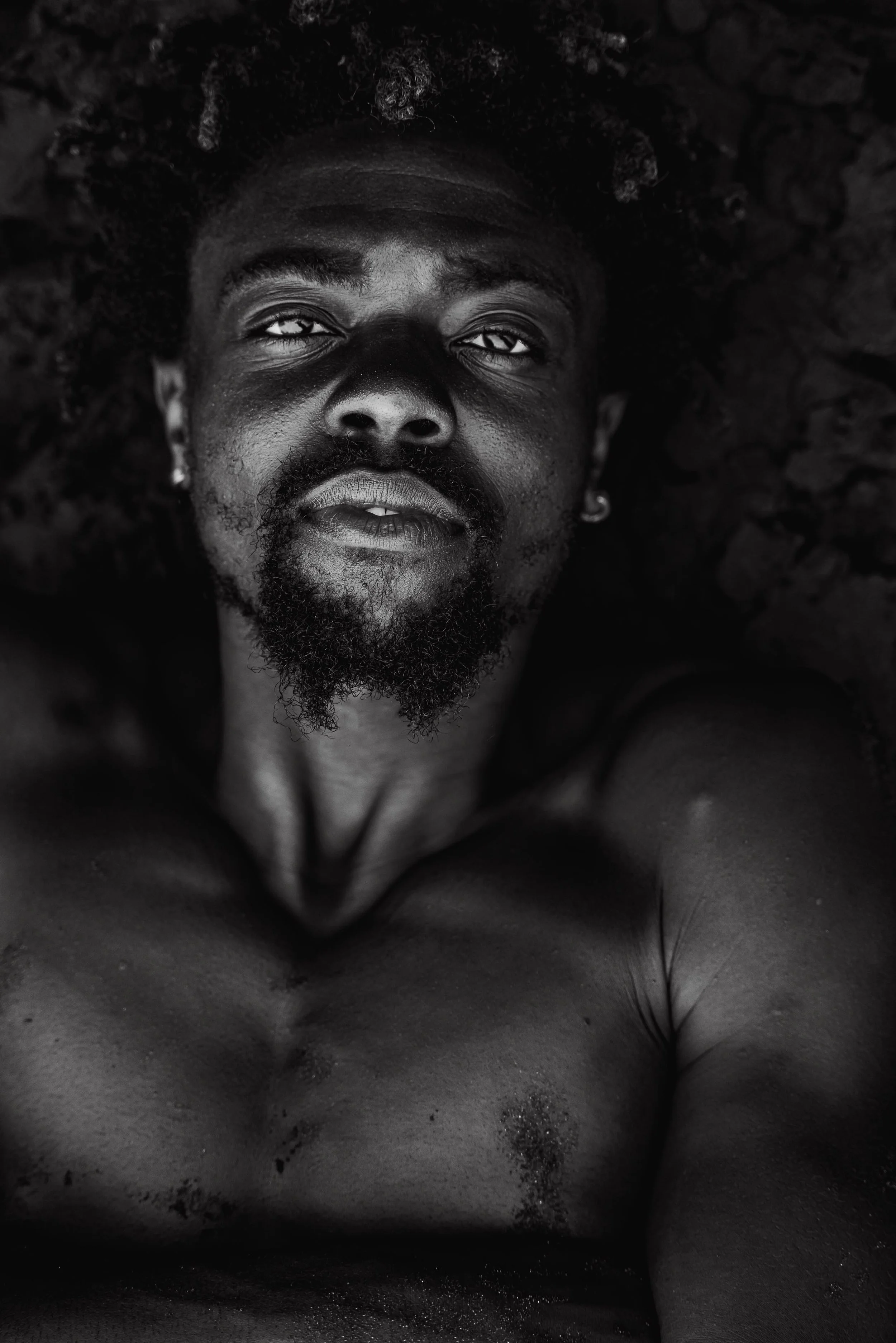 A black and white close-up photo of a shirtless man with facial hair, tattoos, and earrings, lying down and looking into the camera.