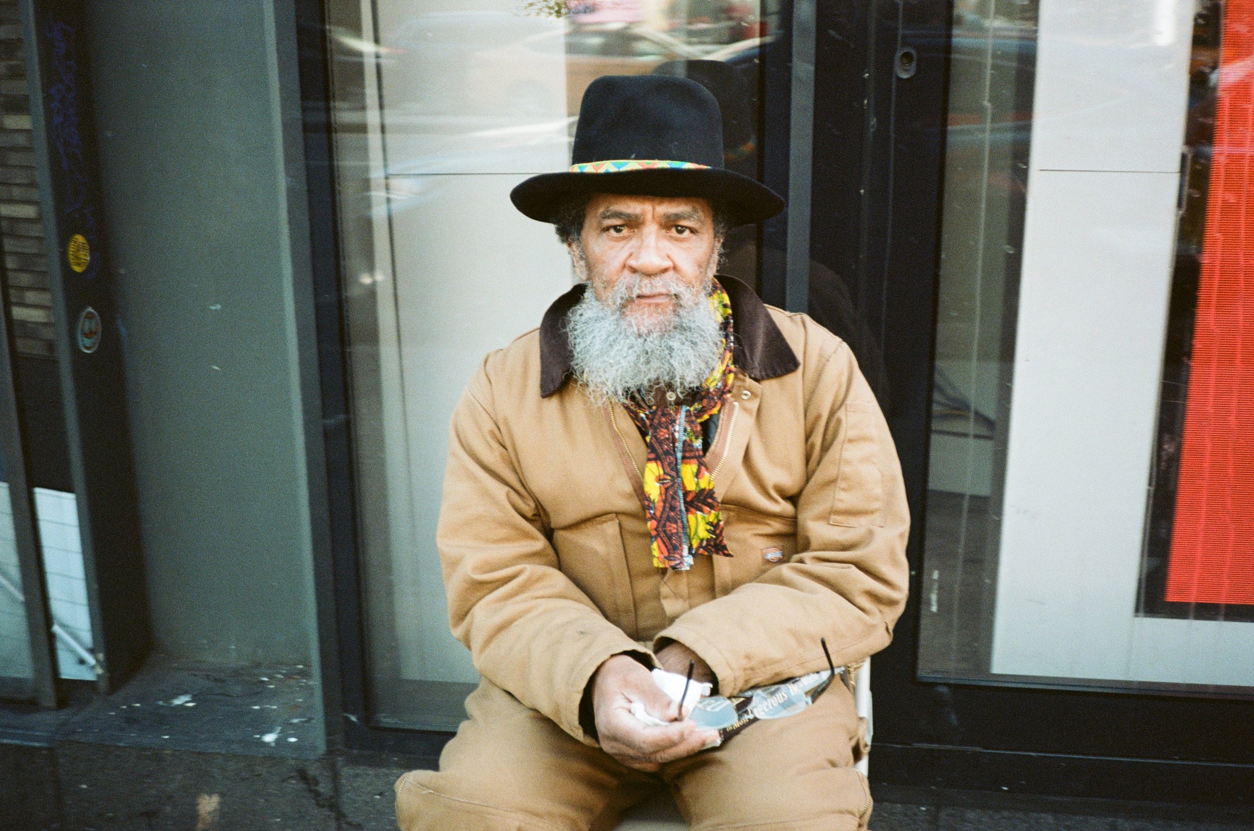 An older man with a gray beard and mustache sitting on a sidewalk in front of a glass building. He wears a black hat with colorful trim, a tan jacket, patterned shirt, and holds sunglasses and a smartphone.