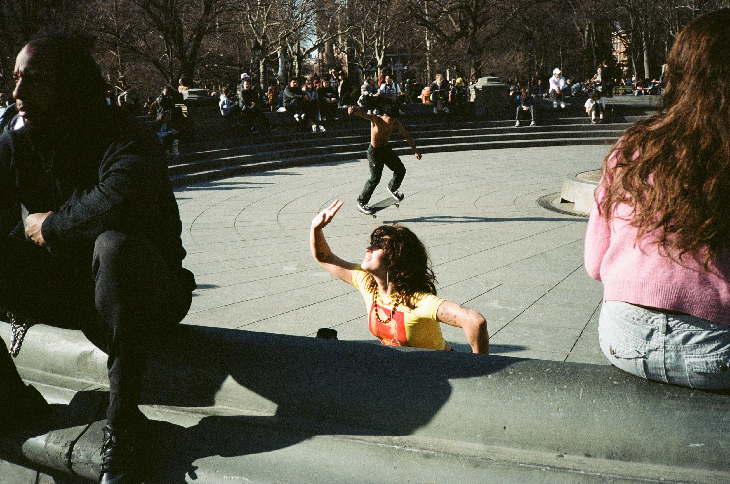 A woman with curly hair, wearing a yellow shirt and red overalls, is sitting on a ledge and waving. In the background, a shirtless skateboarder is skating on a circular paved area, with people sitting on steps and walking nearby.