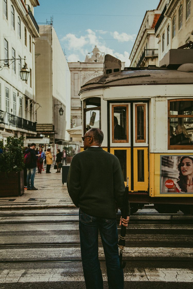 A man holding an umbrella stands at a crosswalk in a city street with a yellow and white tram passing by and several pedestrians walking on the sidewalk.