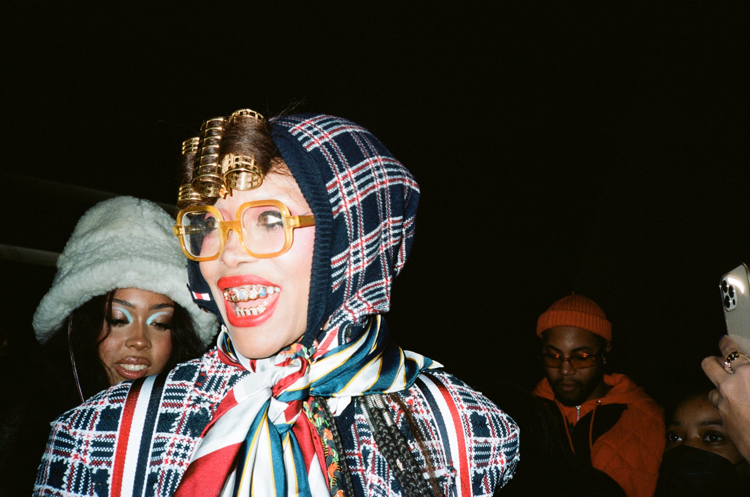 Woman with blonde hair styled in curlers, wearing large yellow glasses, a plaid headscarf, and a colorful patterned coat, smiling and sticking out her tongue at night. In the background, there are people wearing hats and jackets.