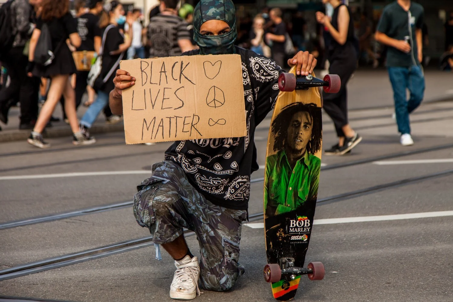 A protester on a skateboard kneeling on the street, holding a cardboard sign that says 'Black Lives Matter' with a heart, peace sign, and infinity symbol, wearing a face covering and patterned clothing. The skateboard has a Bob Marley image. People a