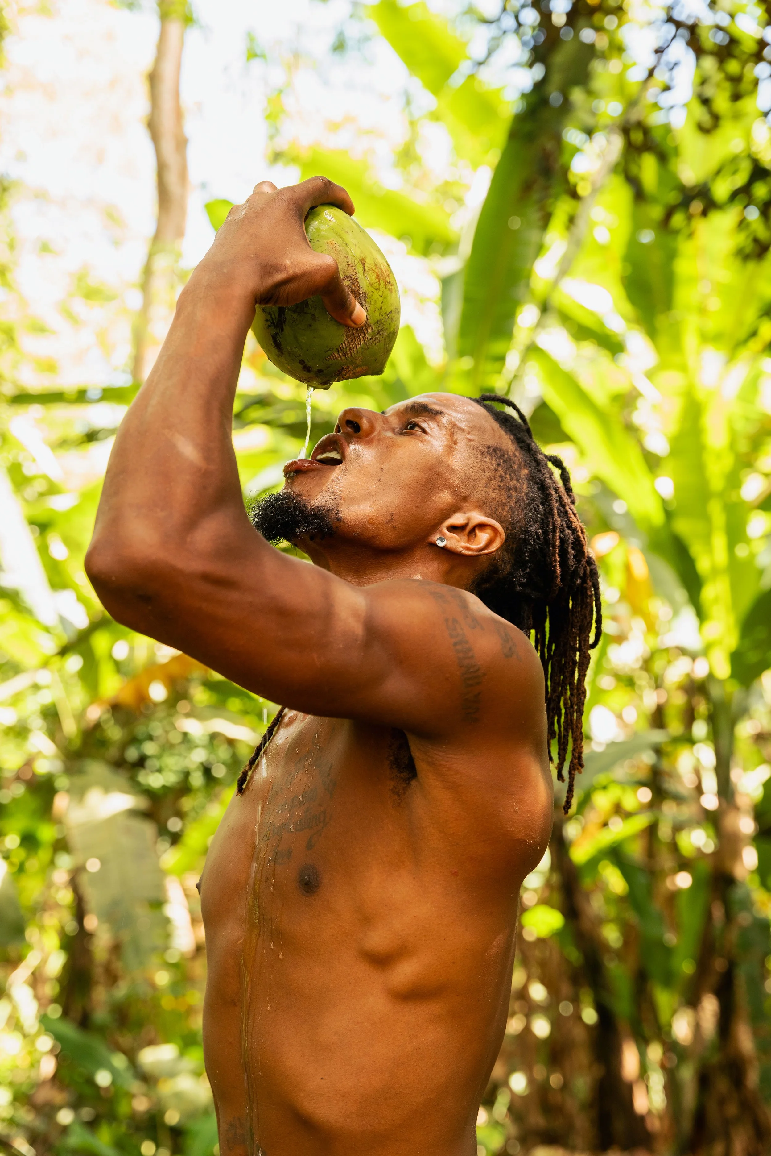 A shirtless man with natural locs and tattoos is pouring coconut water from a green coconut into his mouth in a lush jungle environment.