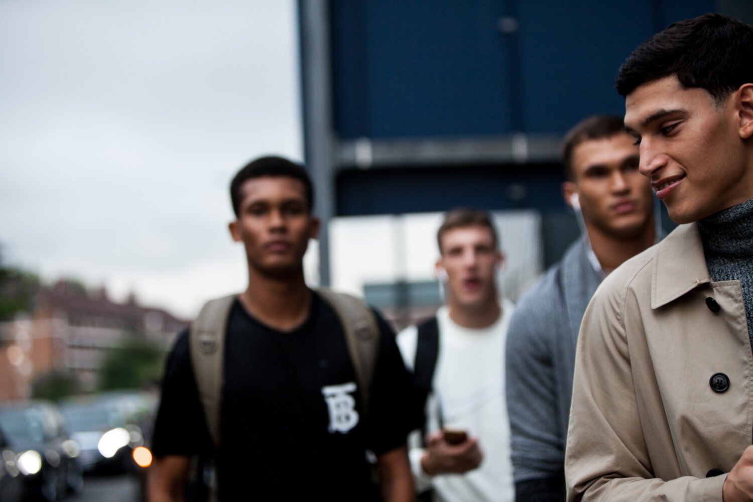 A group of four young men standing outdoors, with one in the foreground wearing a tan trench coat and smiling, while the others look serious or focused. The background shows blurred cars and buildings.