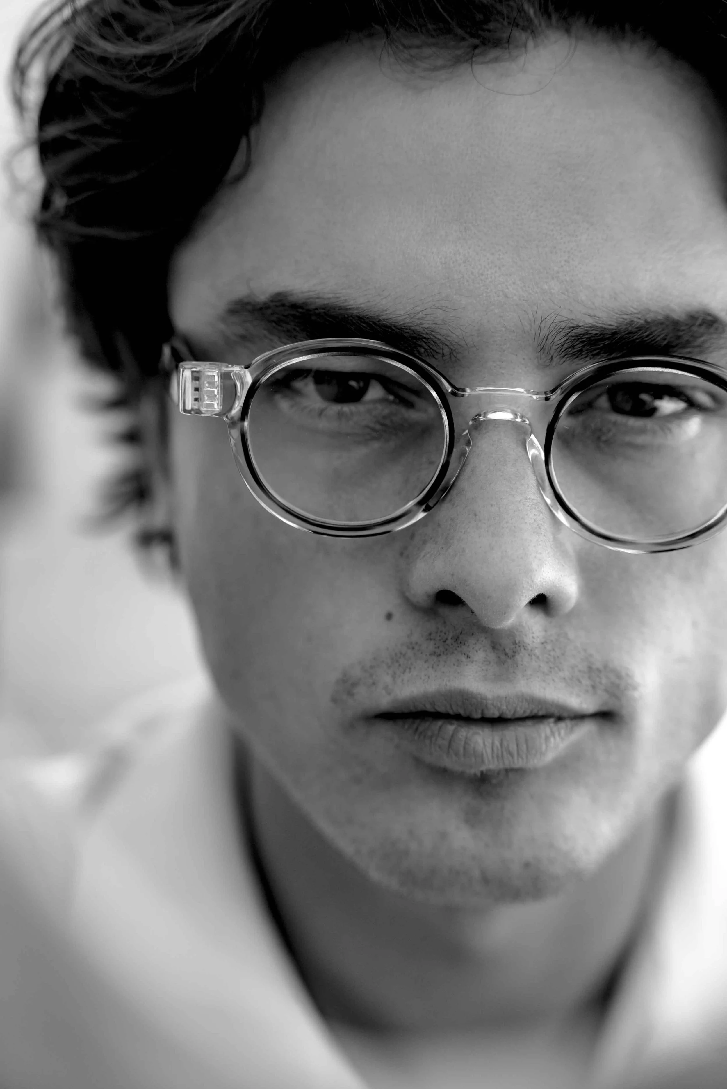 Close-up black and white photo of a young man with dark hair and round glasses, looking slightly downward.
