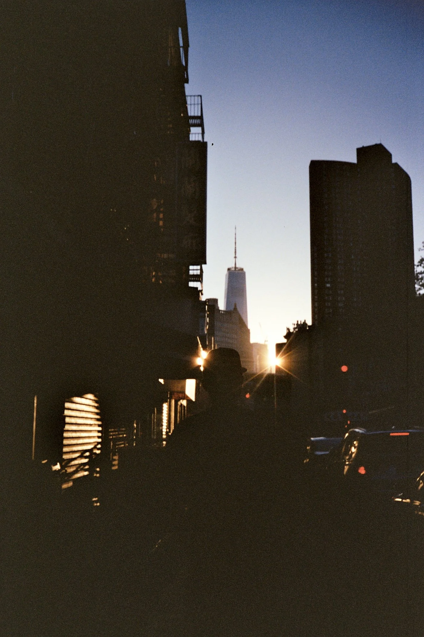 Silhouette of a person wearing a hat standing on a city street at sunset, with the One World Trade Center visible in the background, and sunlight shining between buildings.