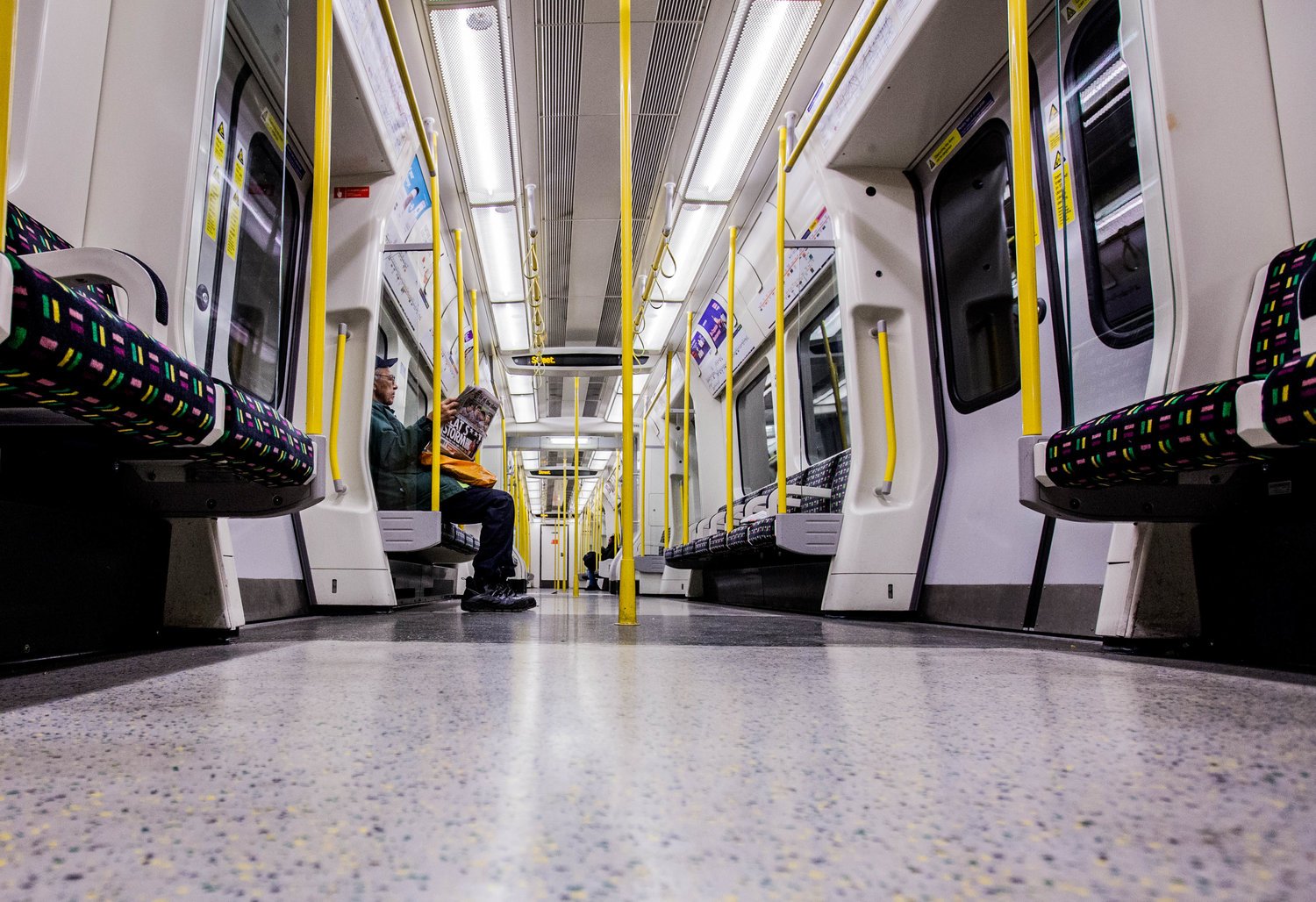 Inside a nearly empty subway train with colorful patterned seats, yellow handrails, and a single passenger reading a newspaper.