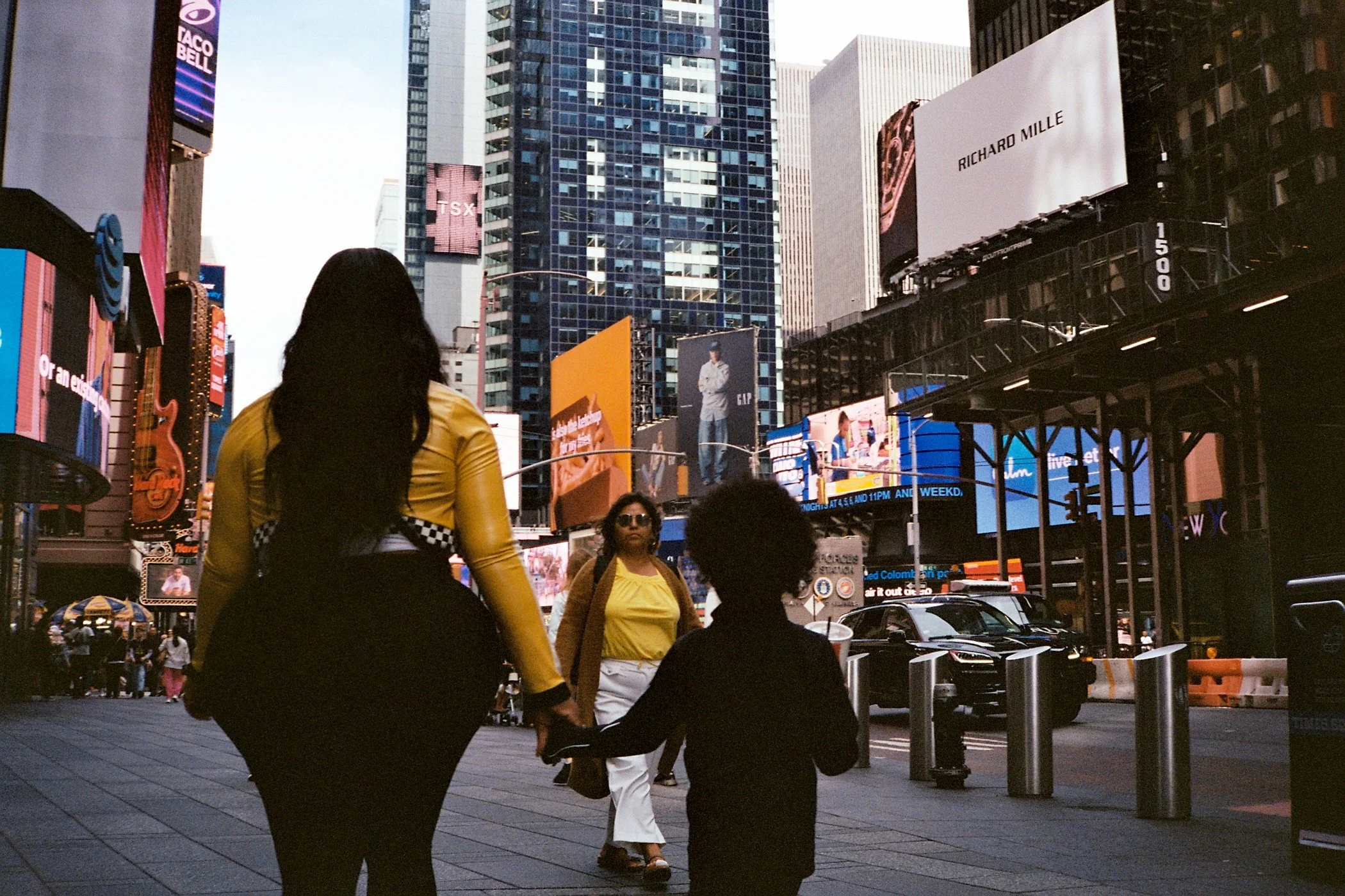 A woman and a young girl holding hands walking in Times Square, New York City, with tall buildings and digital billboards in the background.