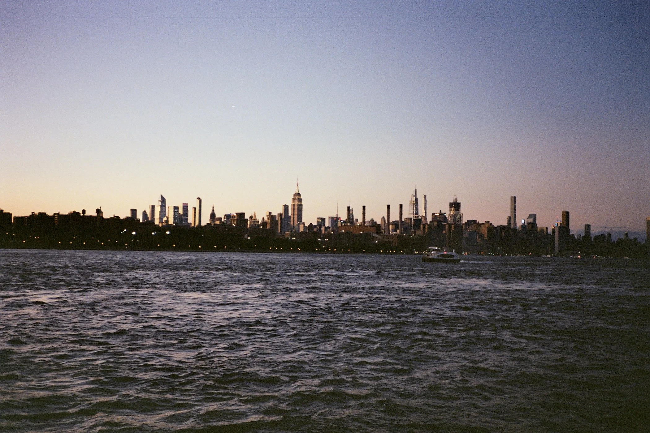 Skyline of Manhattan cityscape at dusk, with water in the foreground and a boat on the river.