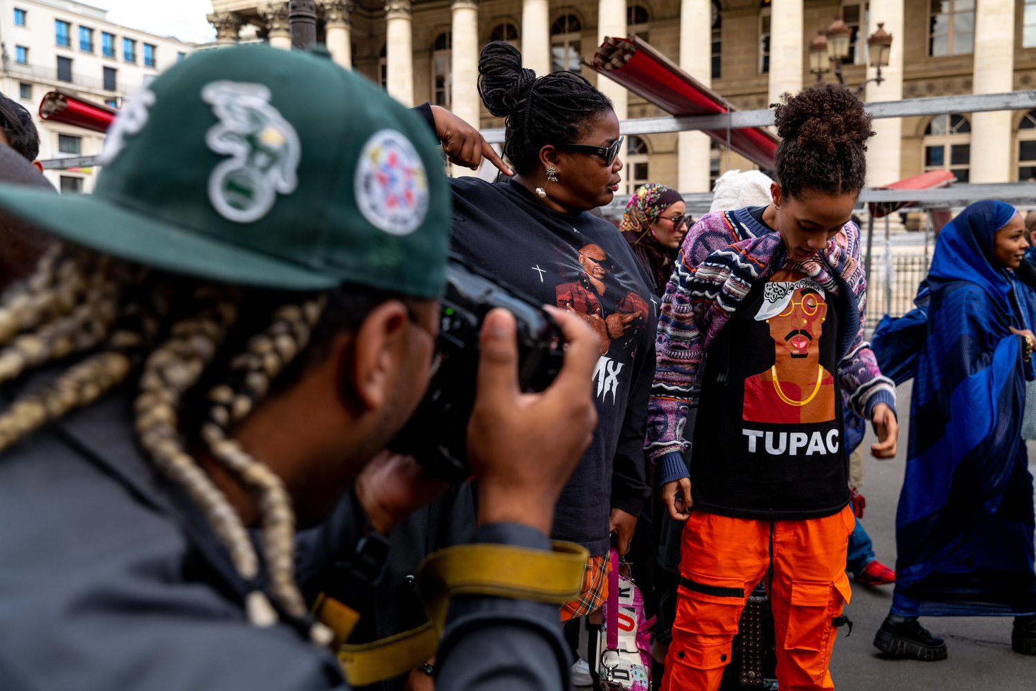 A group of people stand together outdoors, some with their heads bowed, participating in a demonstration or memorial. A woman with glasses and braids is pointing at a young woman wearing a Black Tupac shirt and orange pants. A man with long braided h