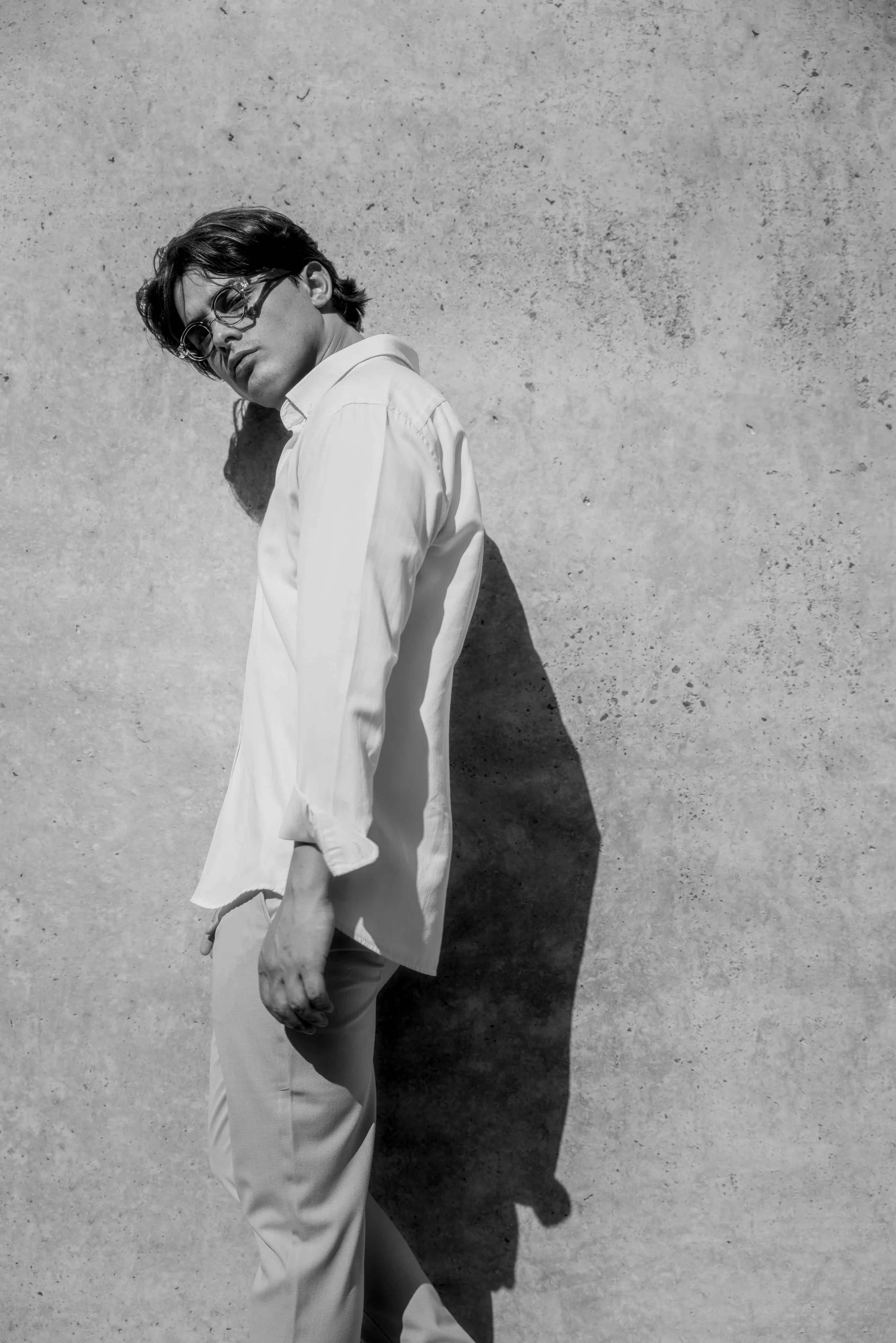 A young man with dark, wavy hair and glasses leaning against a textured concrete wall, dressed in a white shirt and light-colored pants, casting a shadow on the wall.
